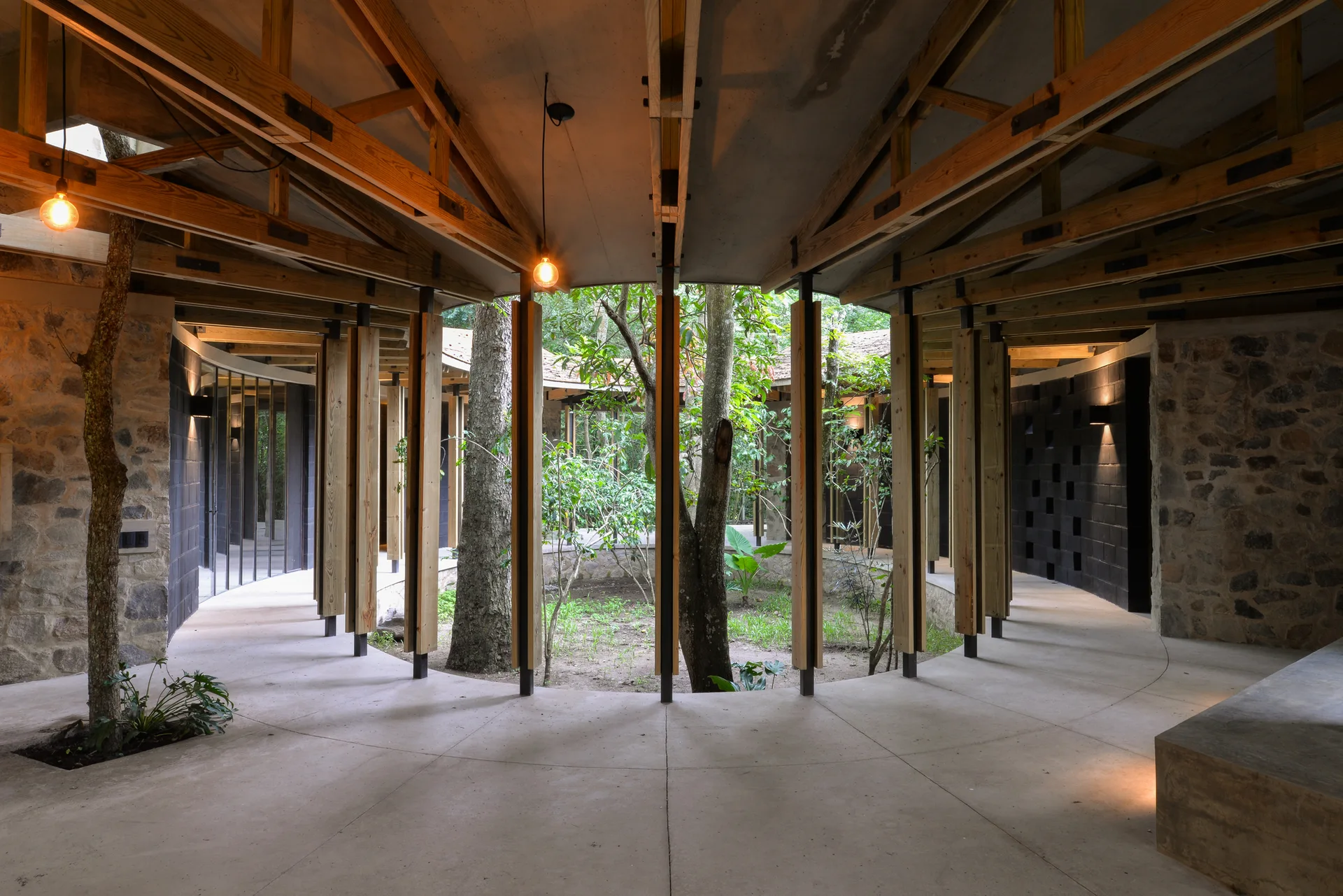 A curved concrete walkway wraps around a central courtyard garden under an exposed timber ceiling structure with hanging bulb pendant lights.