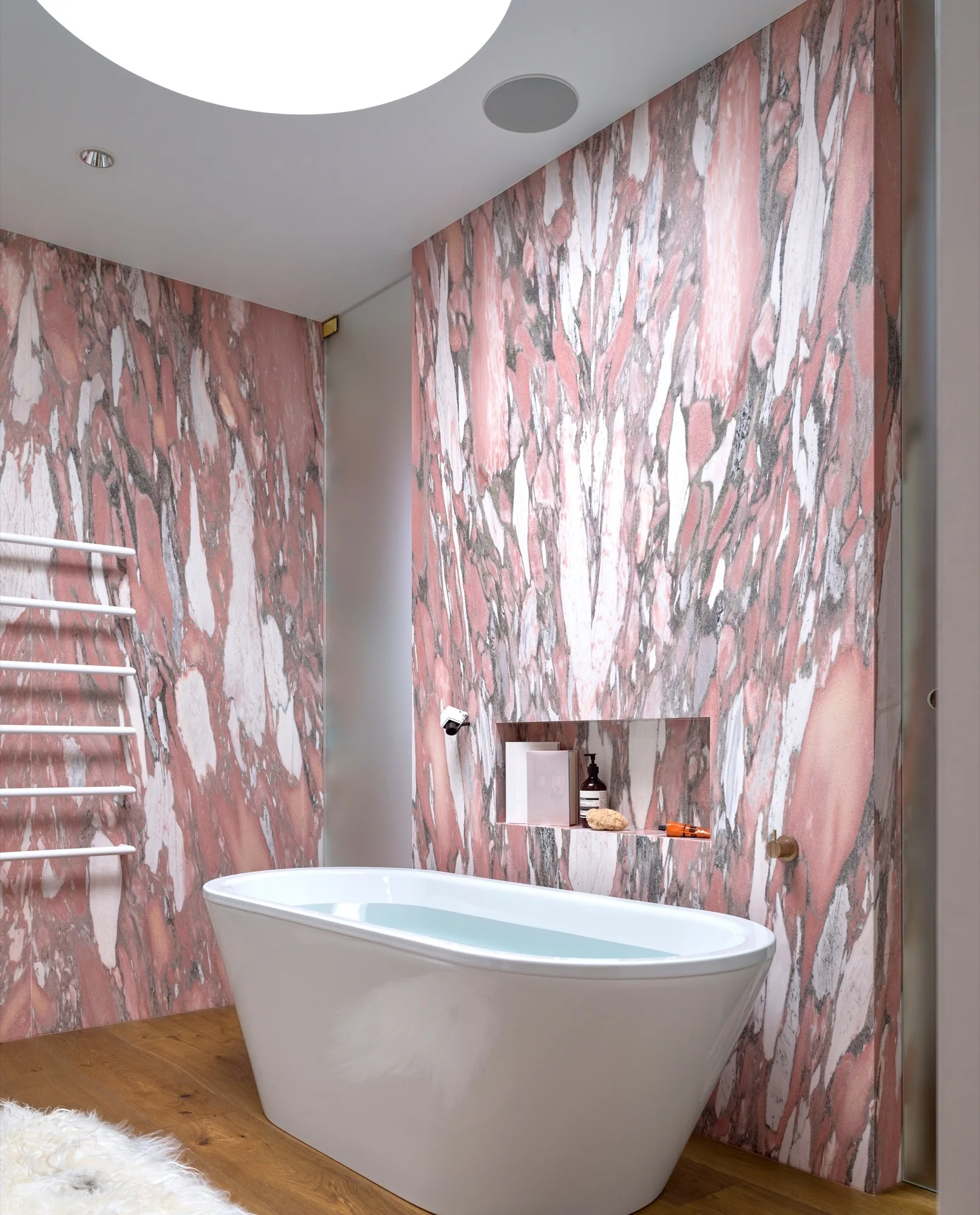 A modern bathroom features a white freestanding bathtub on wood flooring, surrounded by pink veined marble walls and a ladder towel rack.