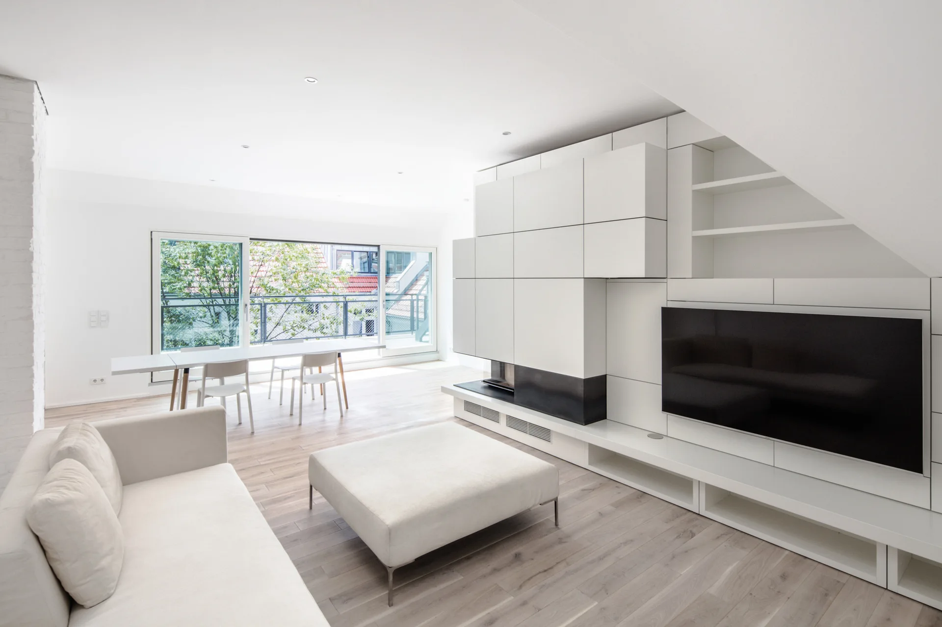 A minimalist white living room features a modern white sofa, a large modular media unit with a television and fireplace, and light wood floors.