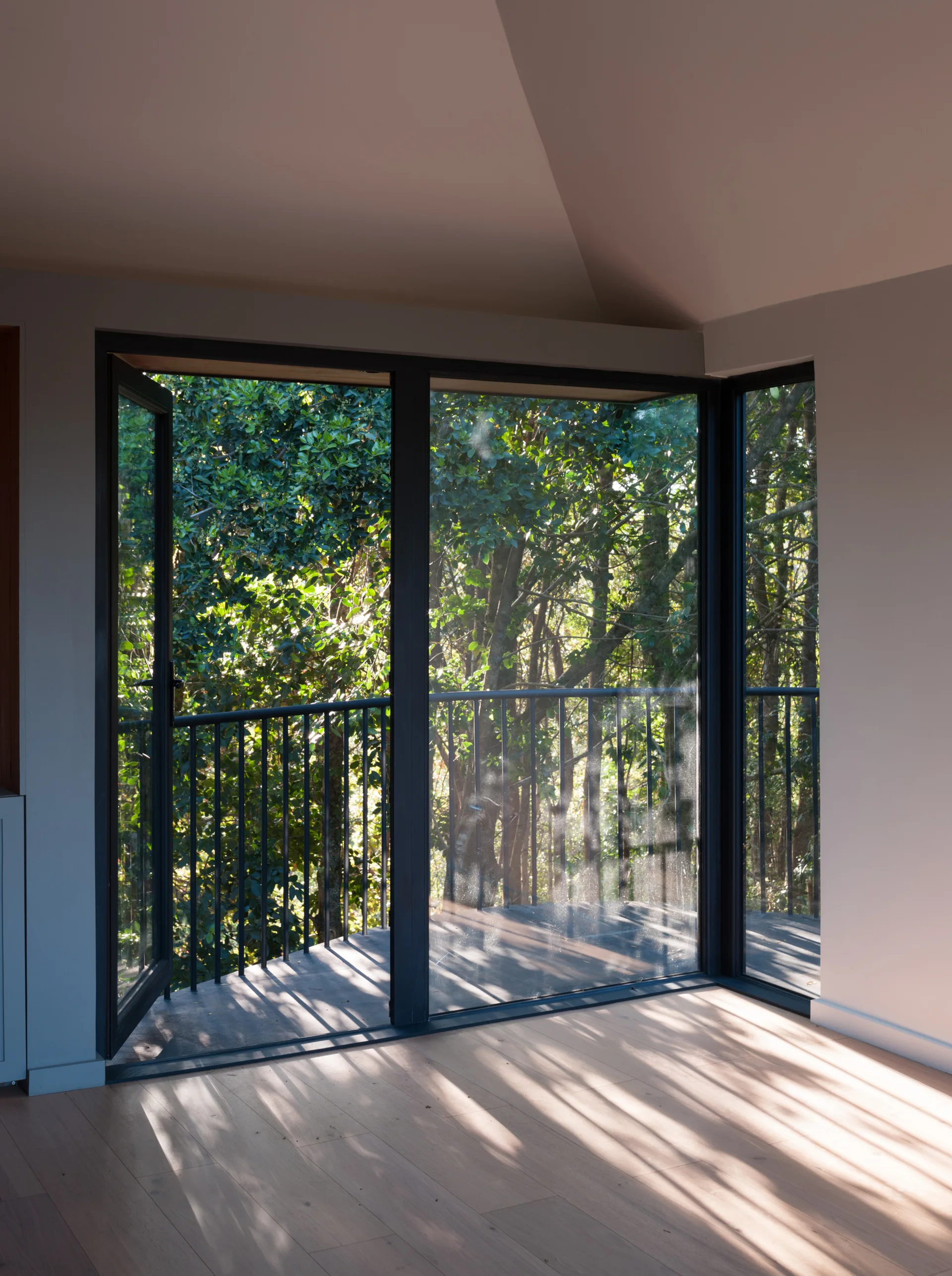 Large black-framed glass corner doors open onto a balcony with a black metal railing, surrounded by lush green forest trees.