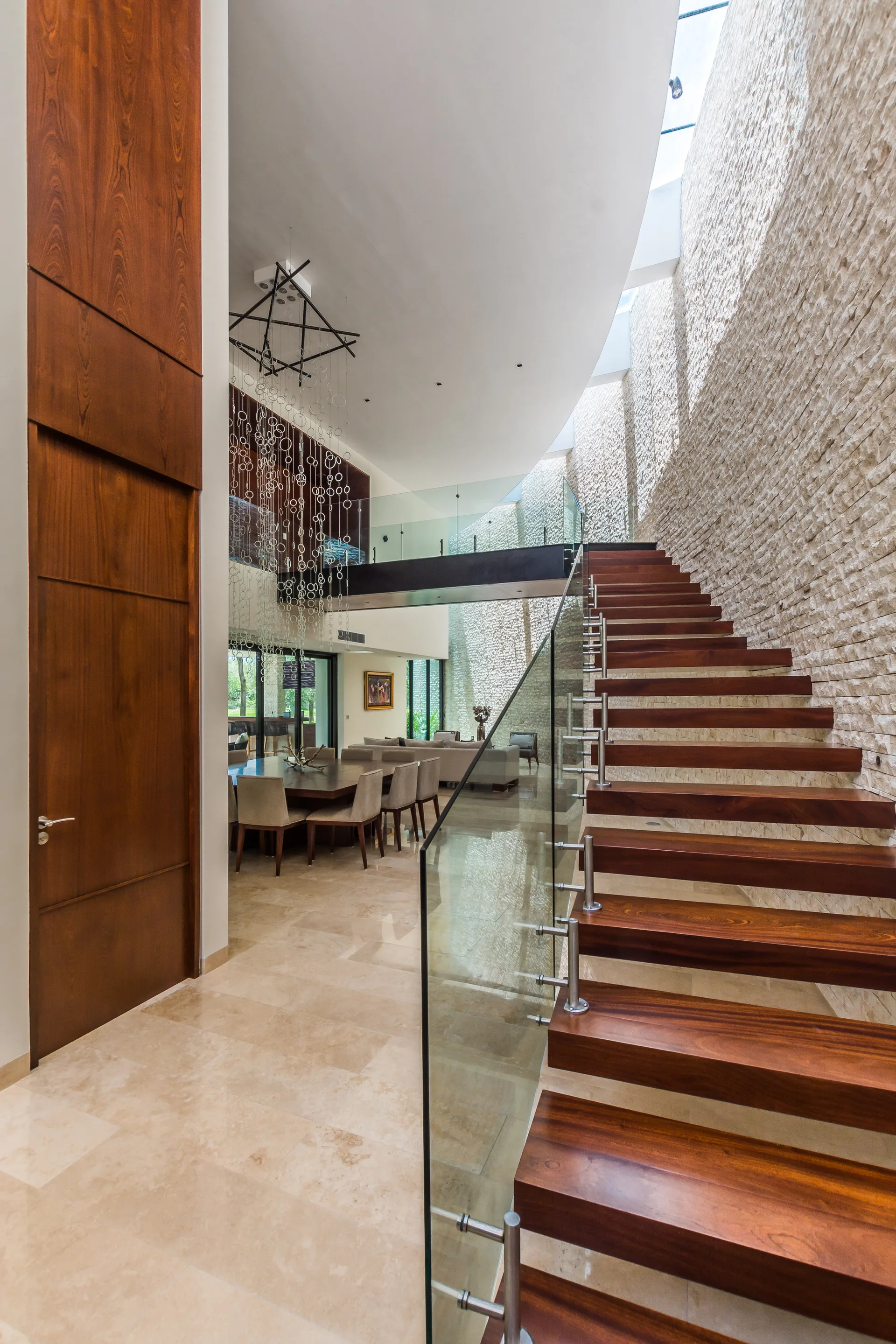 A floating wooden staircase with a glass railing runs alongside a towering textured stone wall under a skylight in a modern open-plan home.