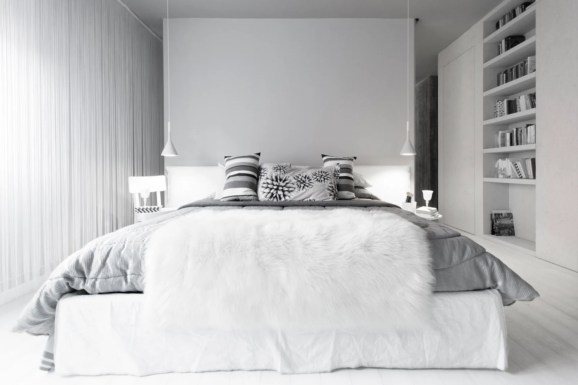 A minimalist white bedroom featuring a bed with grey bedding, a faux fur throw, bedside pendant lights, and a built-in bookshelf.