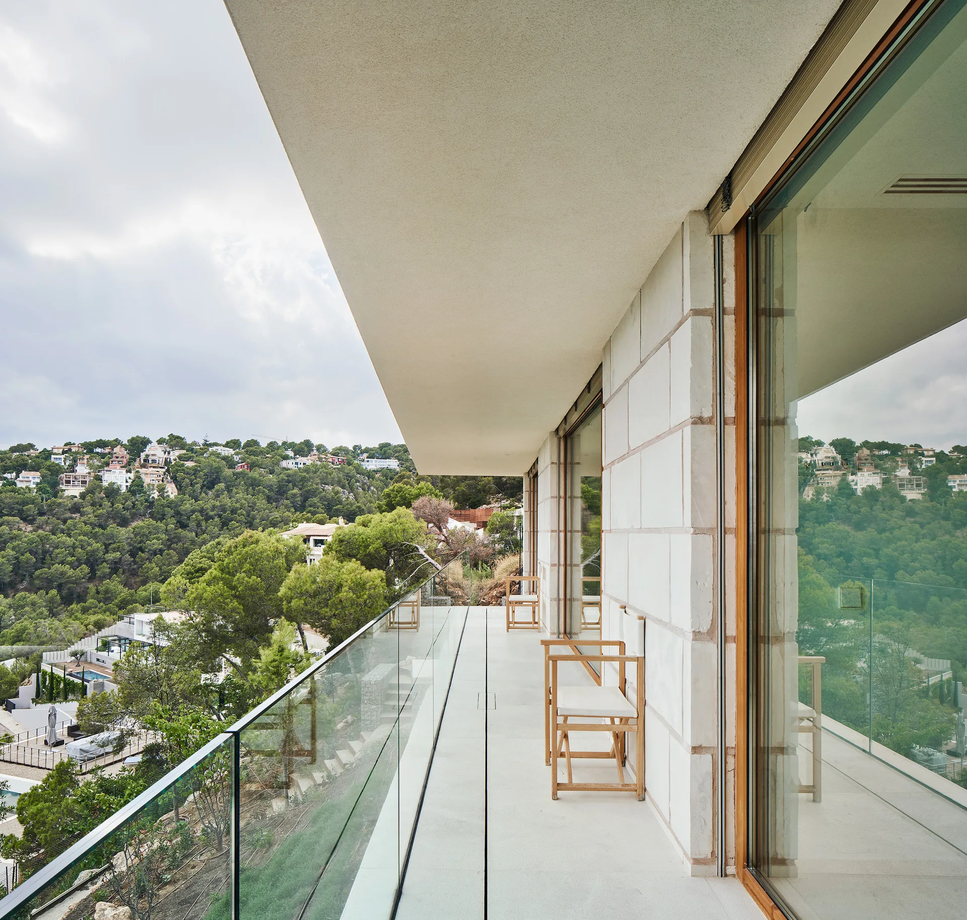 A long modern balcony features light stone cladding, a frameless glass railing, wooden folding chairs, and a scenic hillside view.