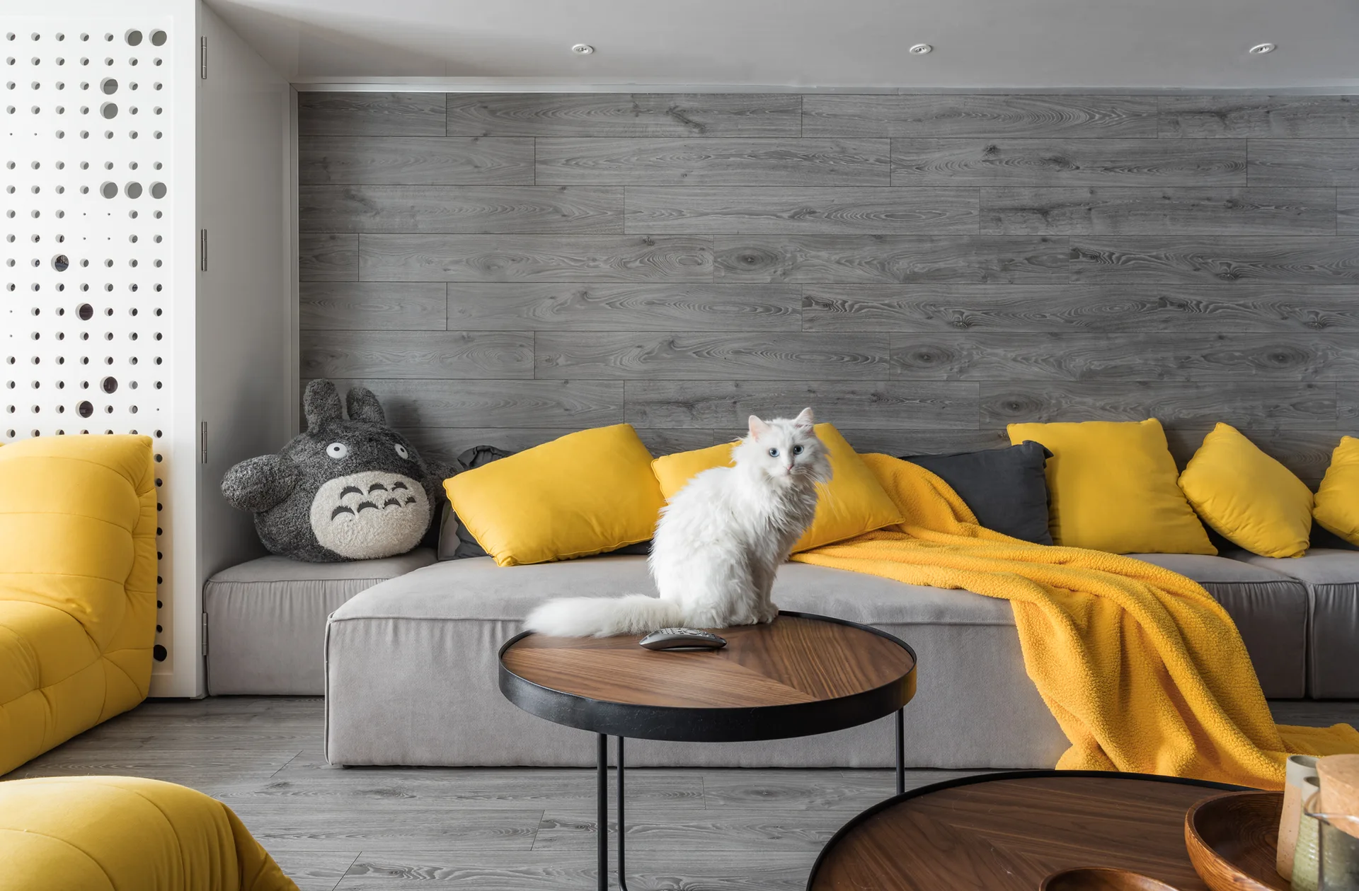 A modern living room features a grey sectional sofa with bright yellow cushions and a blanket against a grey wood plank wall.