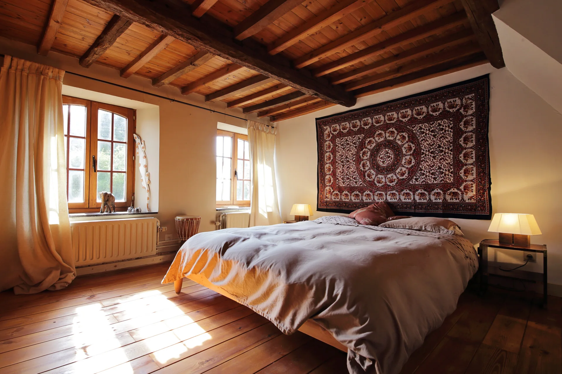 A rustic bedroom features a bed centered under a large patterned wall tapestry, flanked by glowing bedside lamps beneath exposed wood ceiling beams.