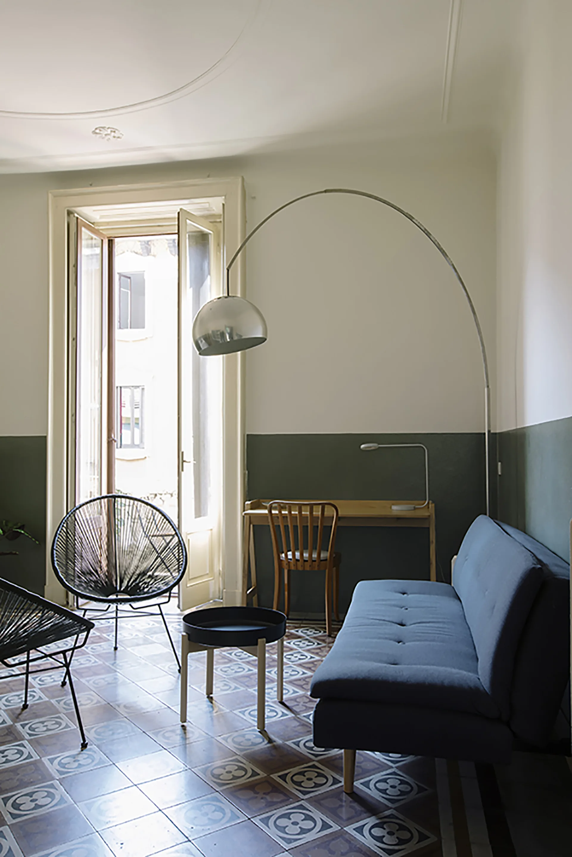 A living area features a blue tufted sofa, a metallic arc floor lamp, a wooden desk, and patterned geometric floor tiles.