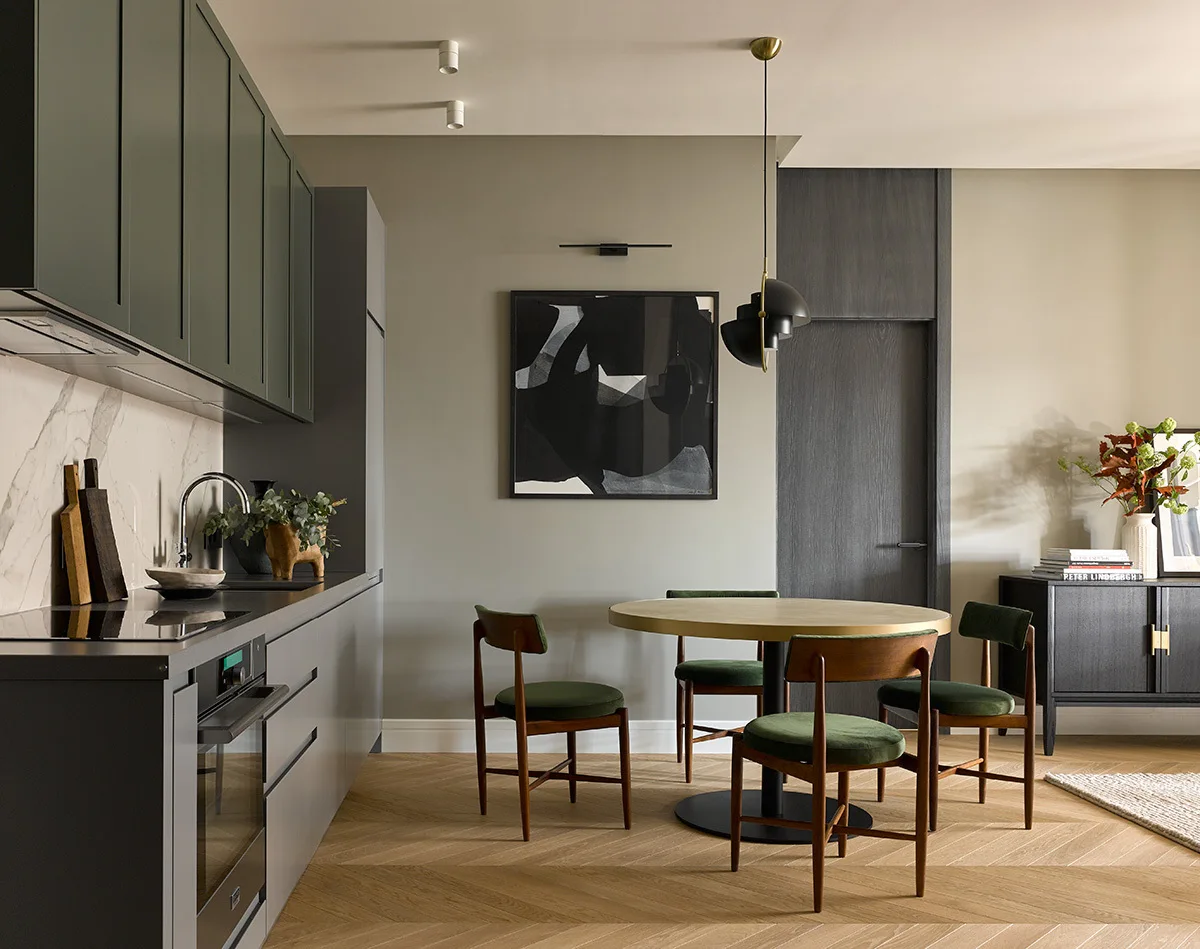 A modern kitchen and dining space with sage green cabinets, a round wooden dining table, and green velvet chairs on herringbone wood floors.