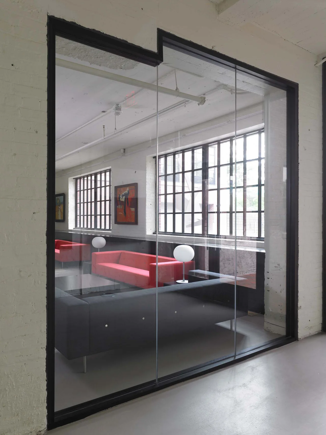 A modern lounge room with red and grey sofas is viewed through a black-framed glass partition wall under an industrial ceiling.