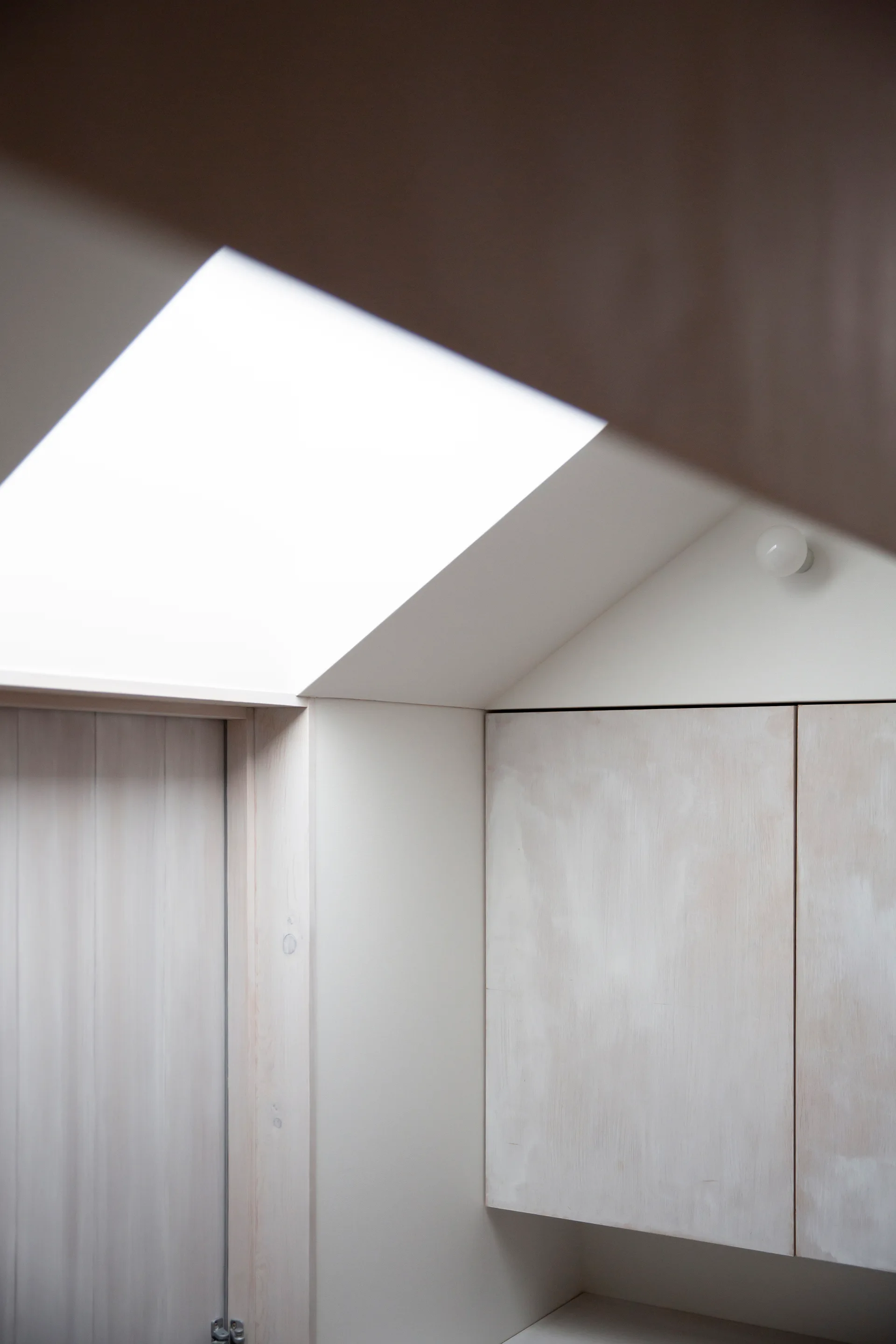 A close-up of built-in limewashed wood cabinets underneath a sloped off-white ceiling featuring a bright skylight.