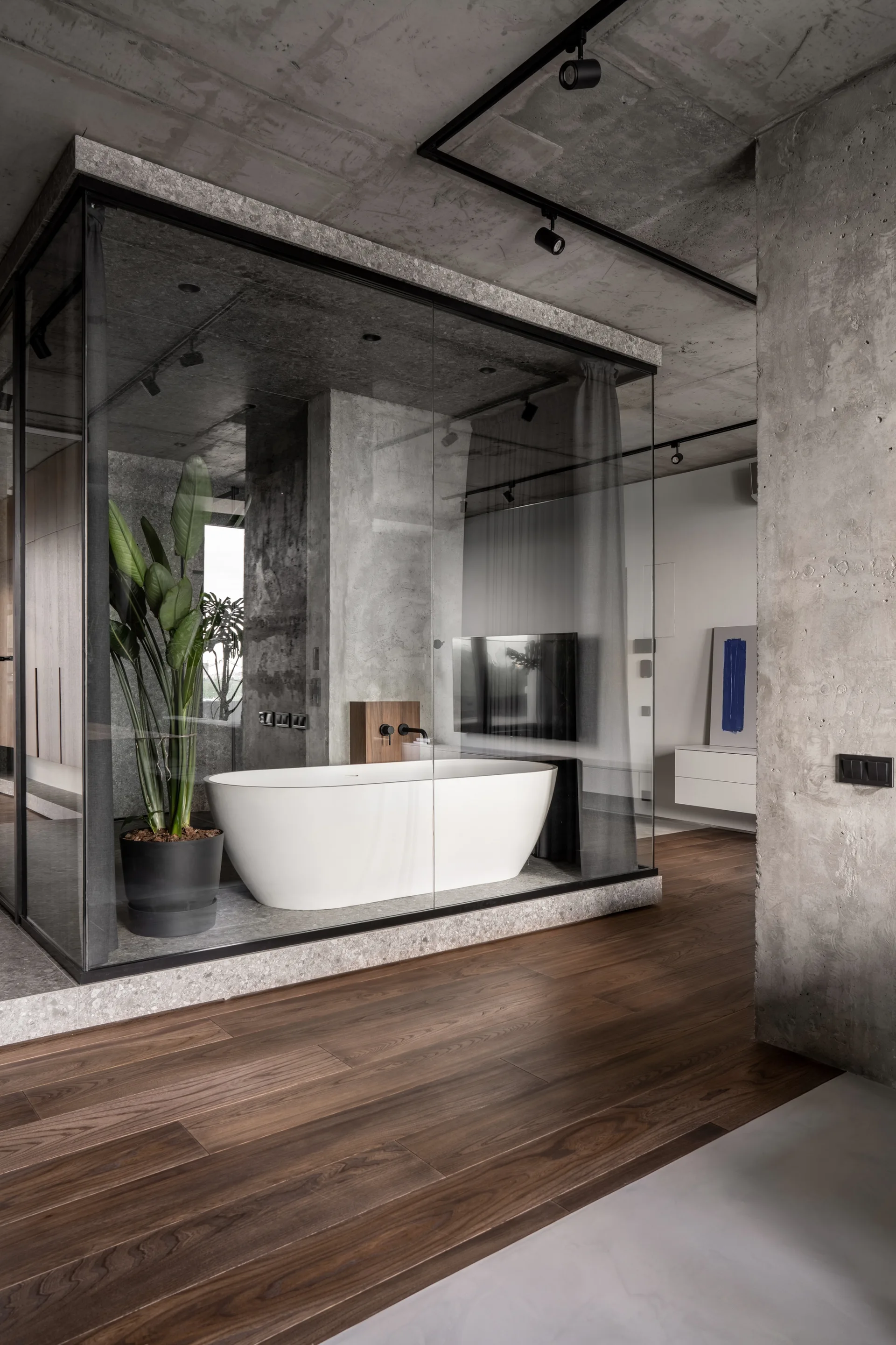 A white freestanding tub is enclosed in a glass cube with raw concrete walls and dark wood floors.
