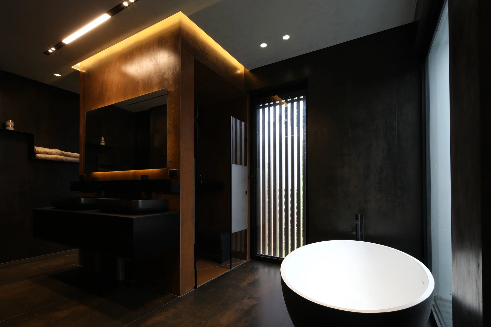 A modern dark bathroom with a white freestanding bathtub, a black floating vanity, a copper-toned feature wall, and warm cove lighting.