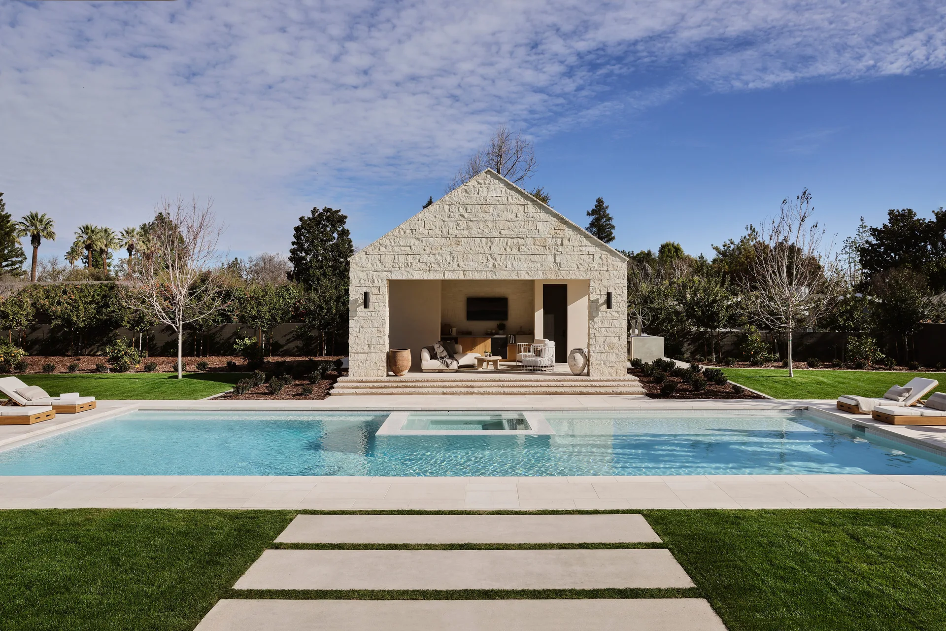 A rectangular swimming pool with clear blue water is flanked by manicured lawns and a gabled, open-front off-white stone pavilion.
