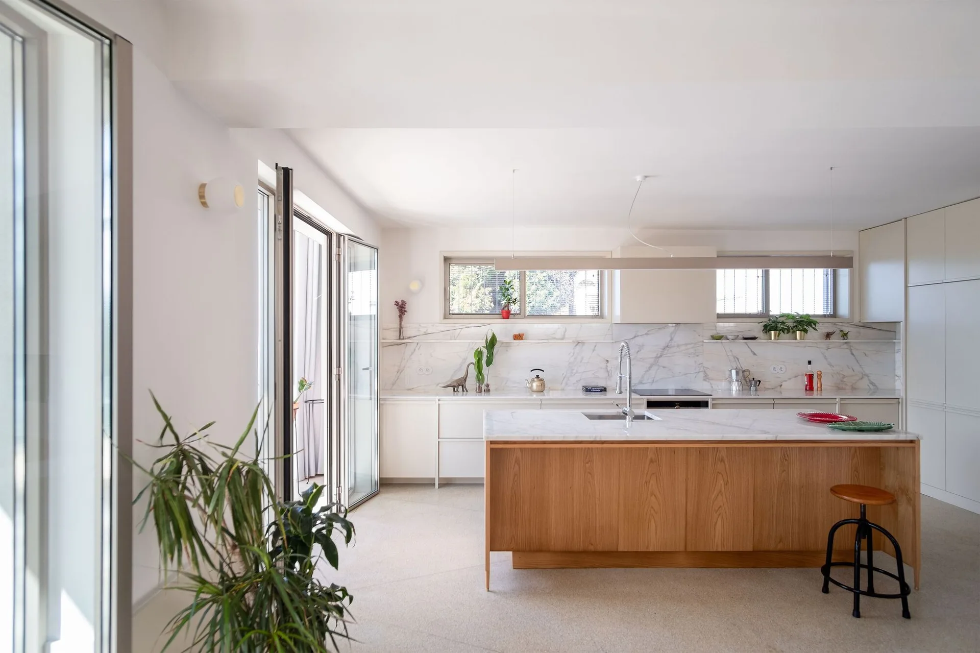 A bright modern kitchen features a wood-toned island with a marble countertop, white cabinetry, a veined marble backsplash, and large glass doors.