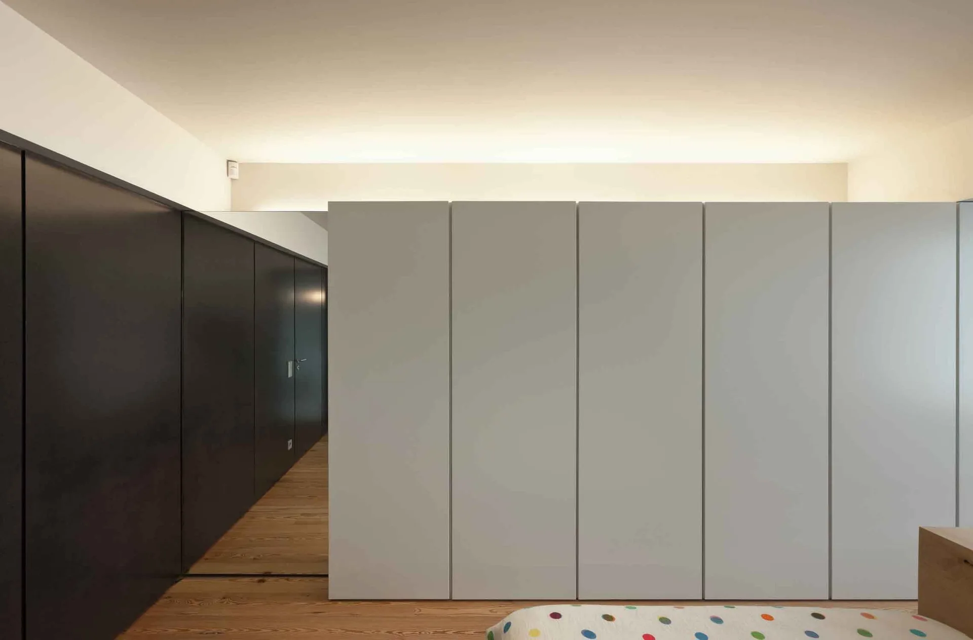 A large built-in white wardrobe sits next to a dark charcoal paneled wall on natural wood plank floors under indirect ceiling lighting.