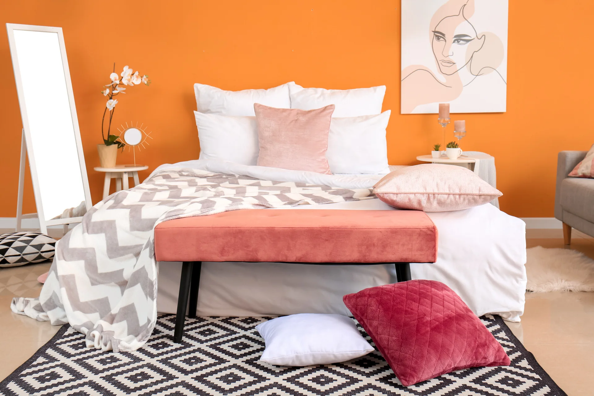A contemporary bedroom features a bold orange accent wall, white bedding, a coral velvet bench, and a black and white geometric rug.