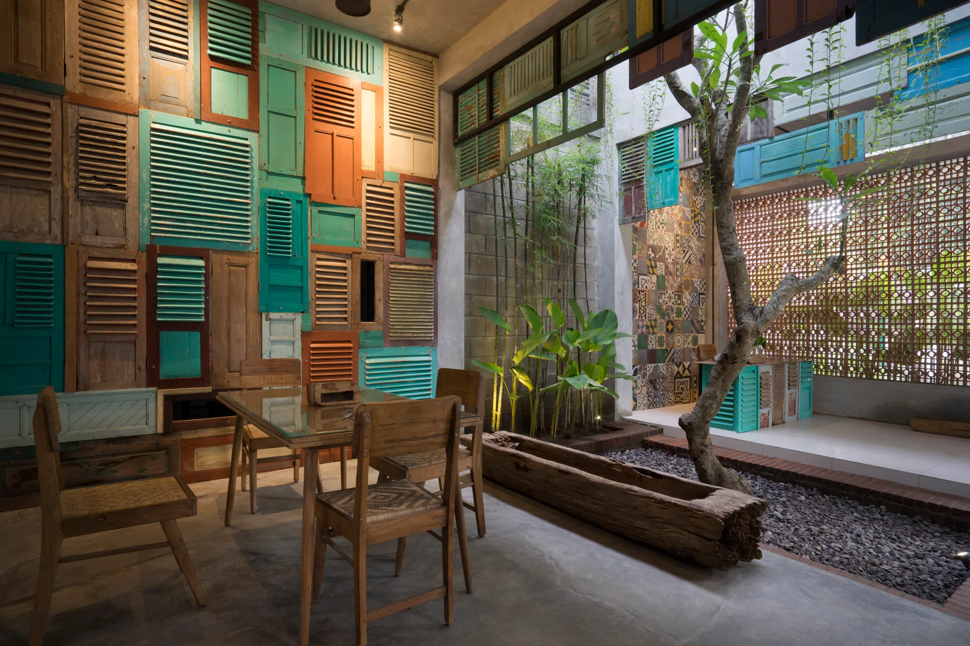A semi-outdoor patio features a wooden dining set next to a wall clad in colorful reclaimed shutters and a garden area with a tree.
