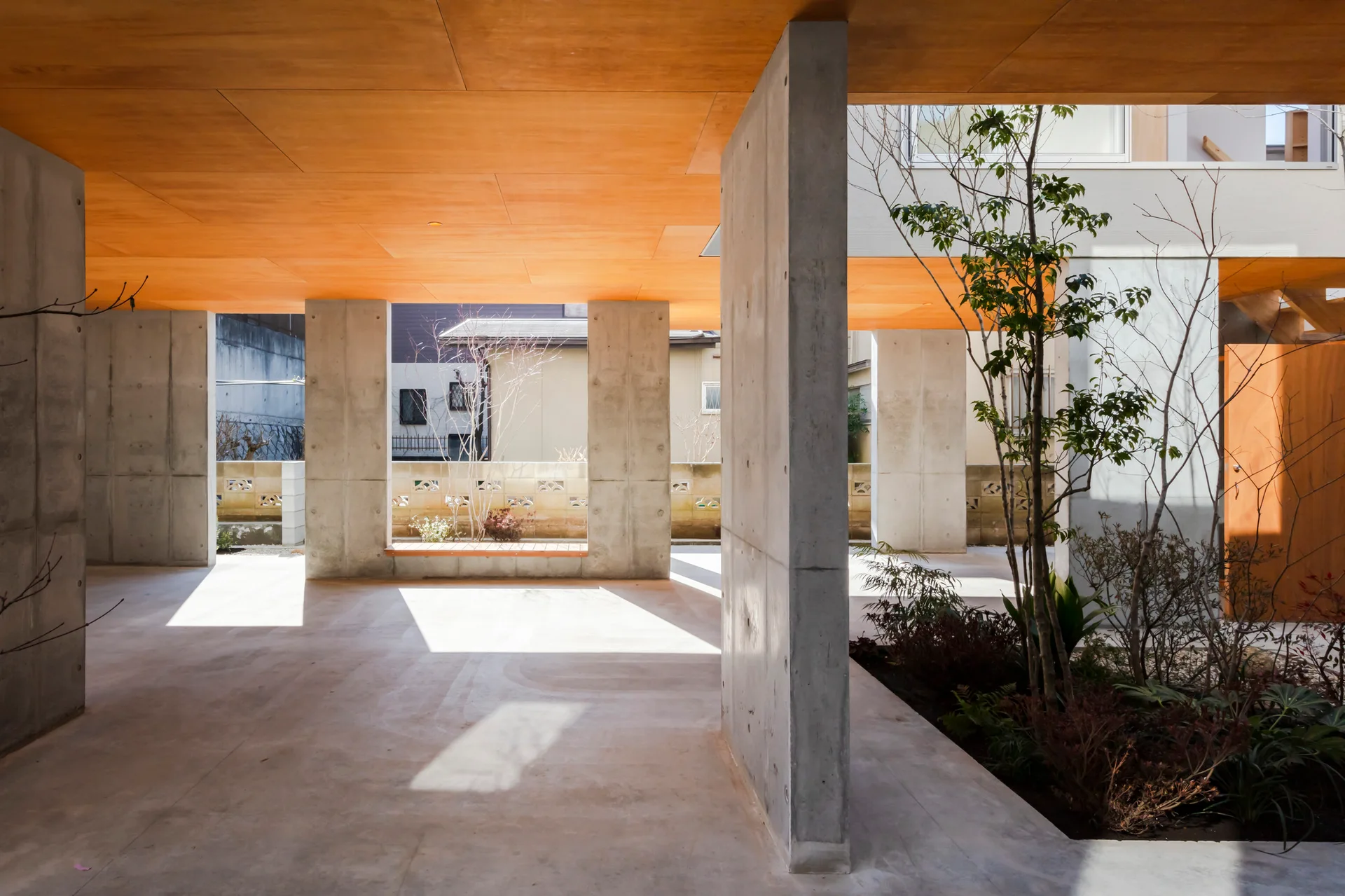 A semi-enclosed modern courtyard featuring robust concrete pillars, a warm wood panel ceiling, and a small landscaped garden bed under natural sunlight.