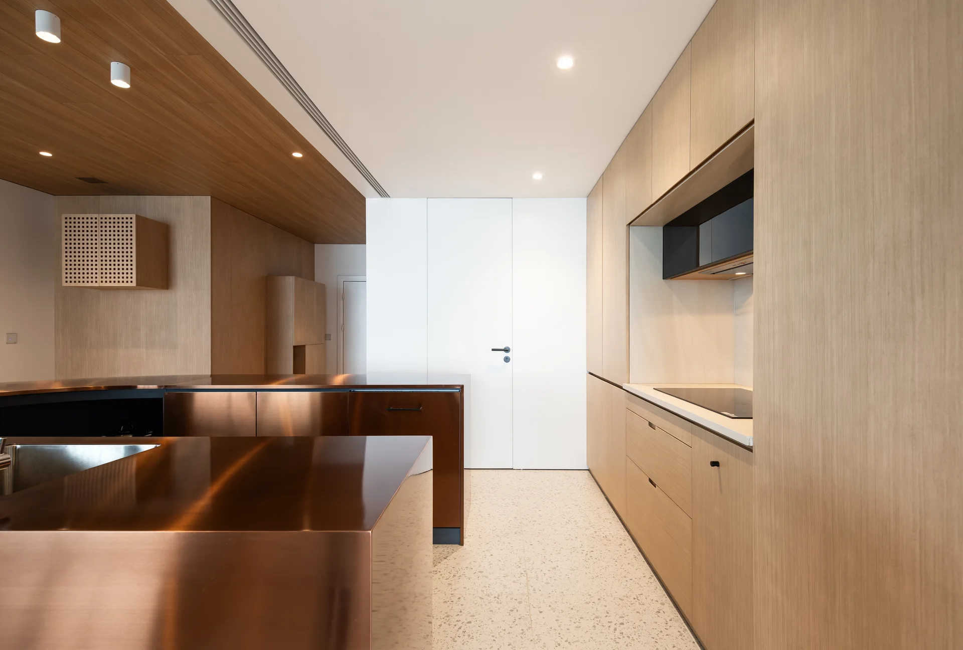 A minimalist kitchen featuring a polished copper-toned island, seamless light wood cabinetry, and speckled terrazzo flooring.