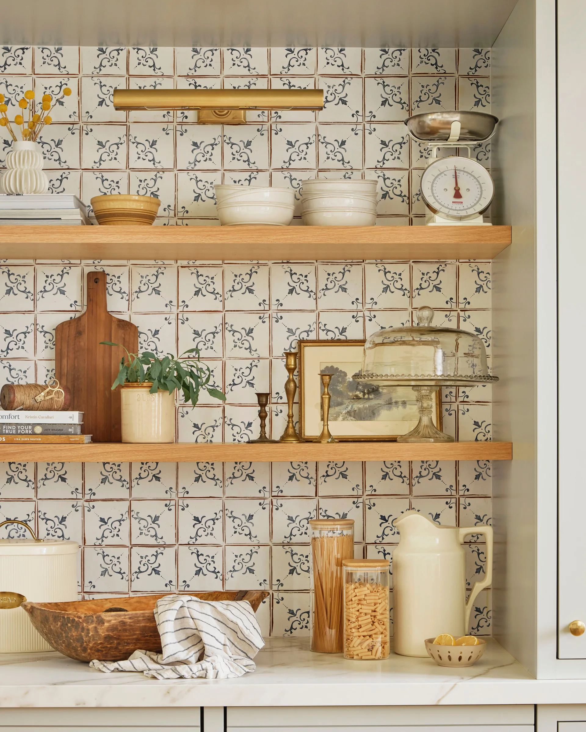 Open wood shelves displaying white ceramic bowls and a vintage scale are mounted on a blue patterned tile wall.
