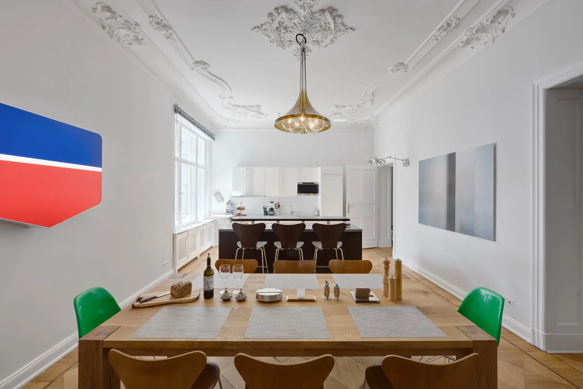 A wooden dining table with mixed wooden and green chairs sits under an ornate plaster ceiling medallion in a bright white open-plan space.