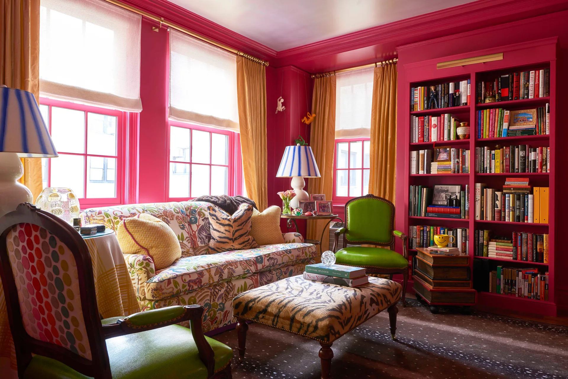A maximalist library featuring glossy hot pink walls and built-in bookshelves, a colorful floral sofa, green armchairs, and a tiger-print ottoman.