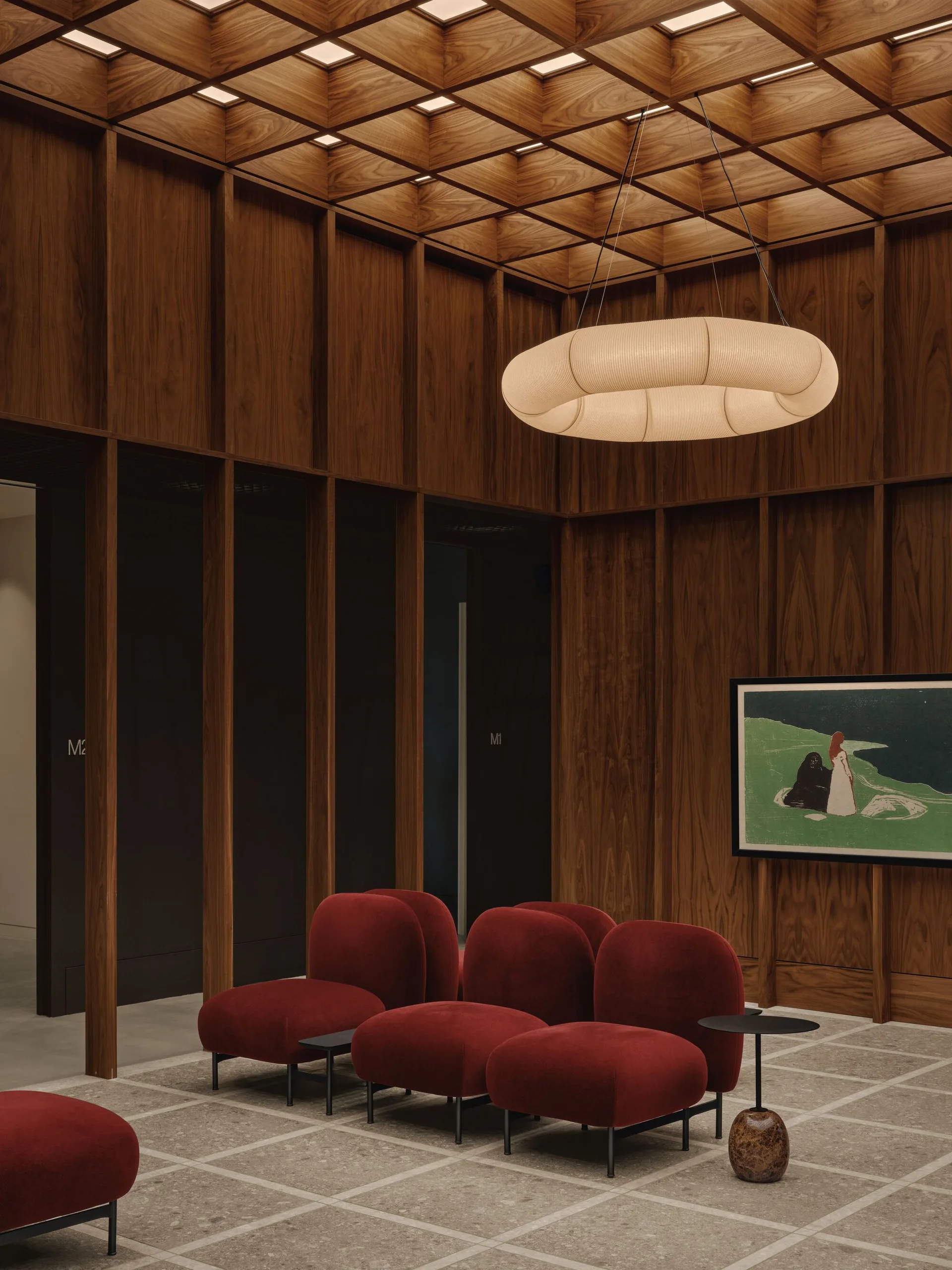 A waiting area with vertical wood paneling, a grid-patterned coffered ceiling, a large hanging ring light, and several low-profile red velvet lounge chairs.