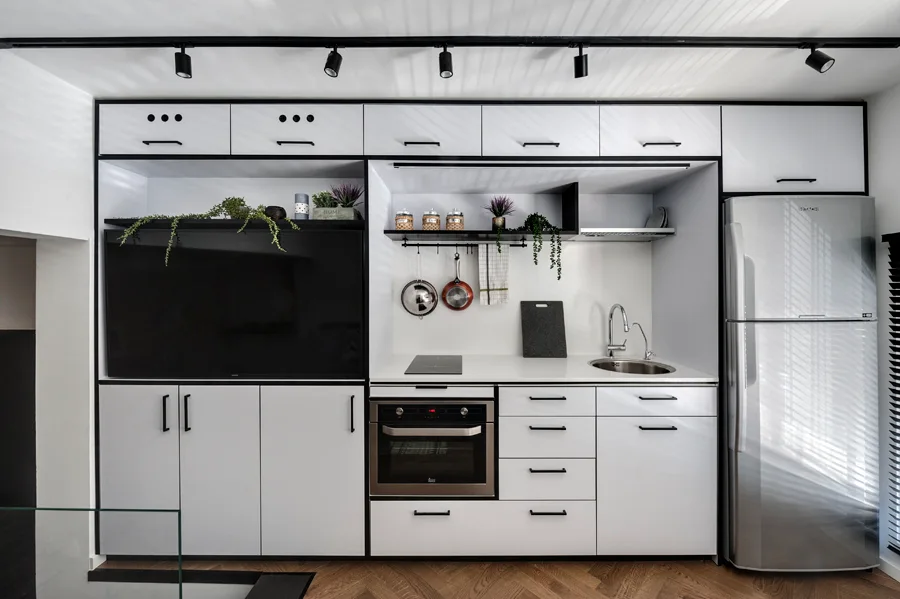 A compact modern kitchenette features white cabinets with black trim, a built-in television, stainless steel refrigerator, and herringbone wood floors.