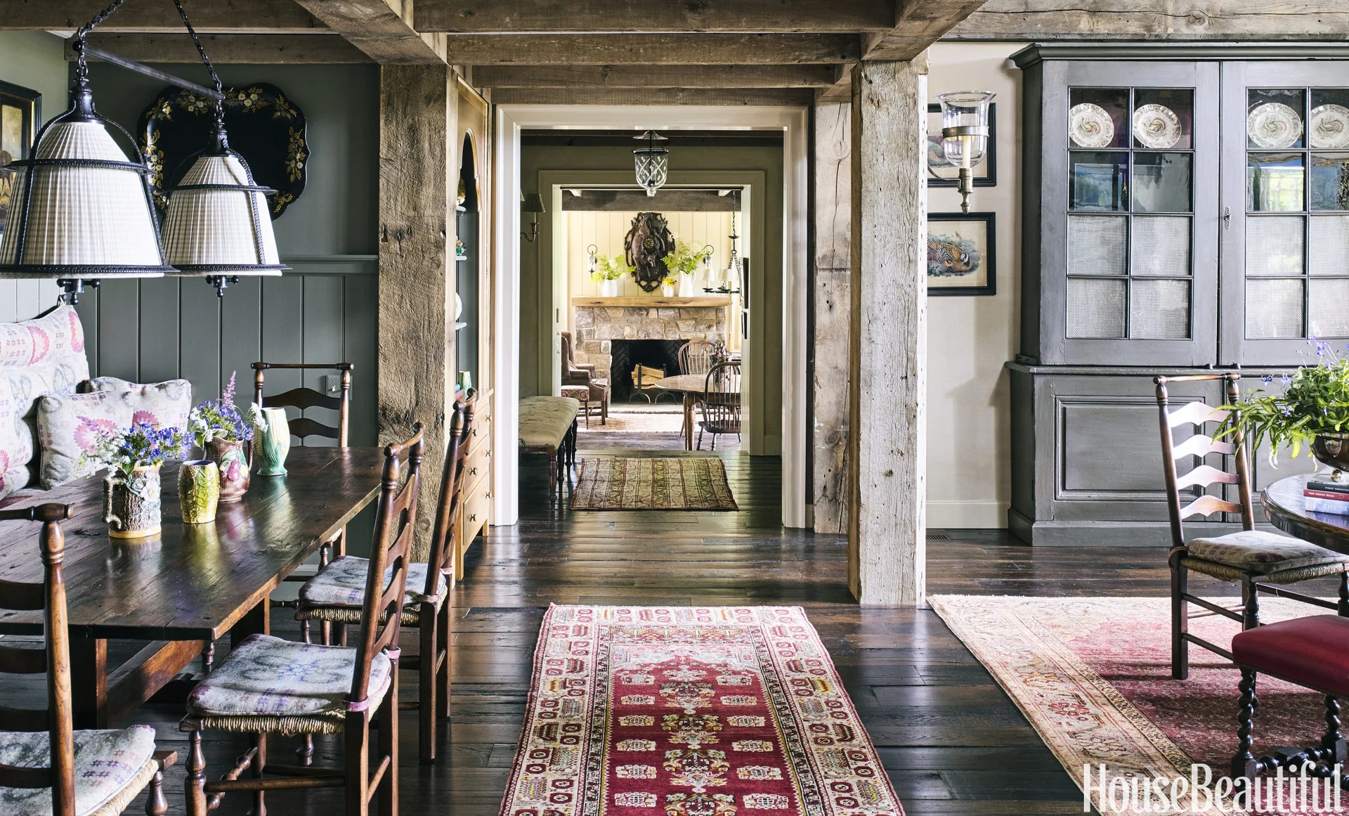 A rustic dining room features a dark wood table with ladder-back chairs, exposed timber posts, patterned rugs, and an open doorway showing a stone fireplace.
