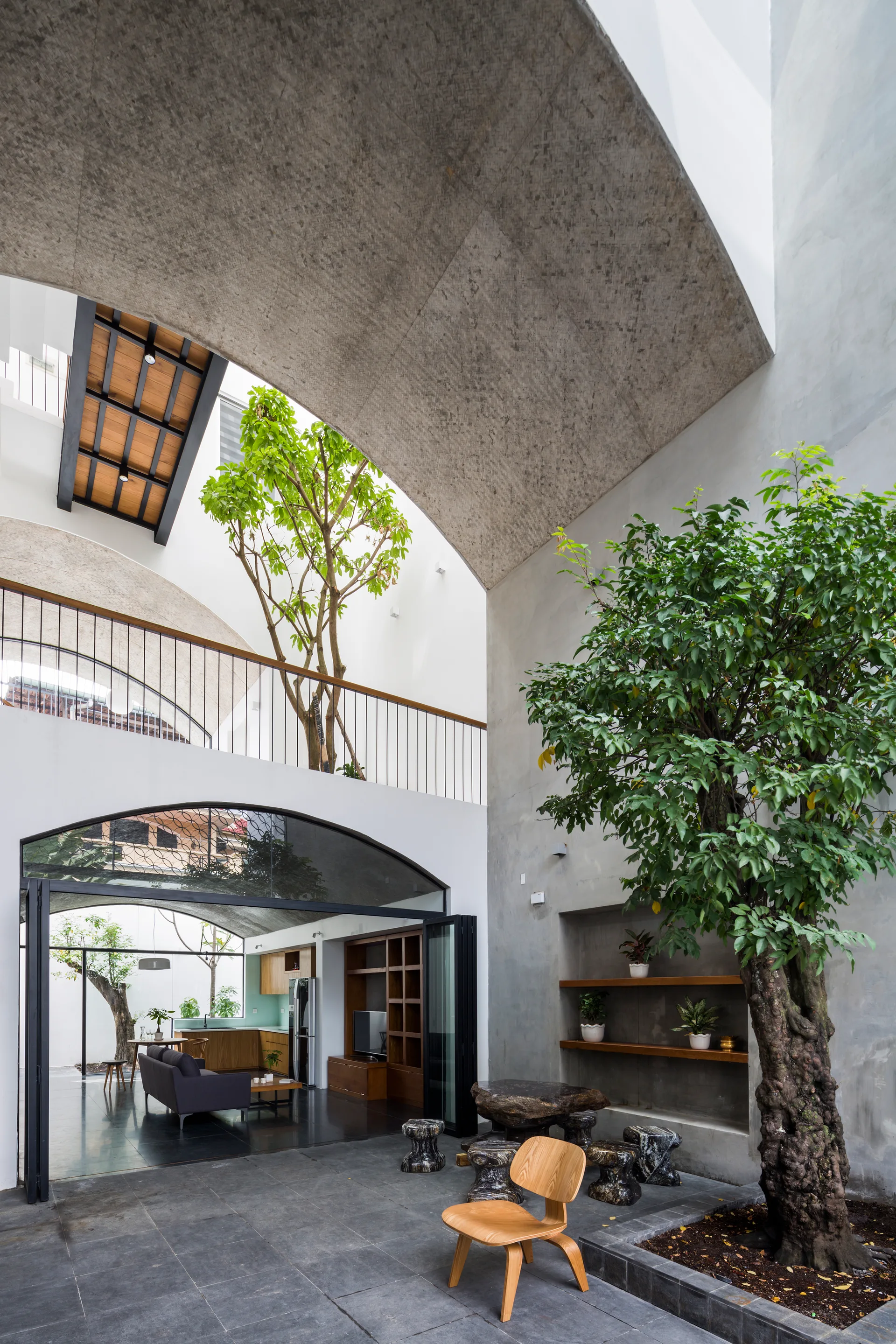 An open courtyard house featuring a large concrete arch, mature indoor trees, and a wooden lounge chair on dark stone flooring.
