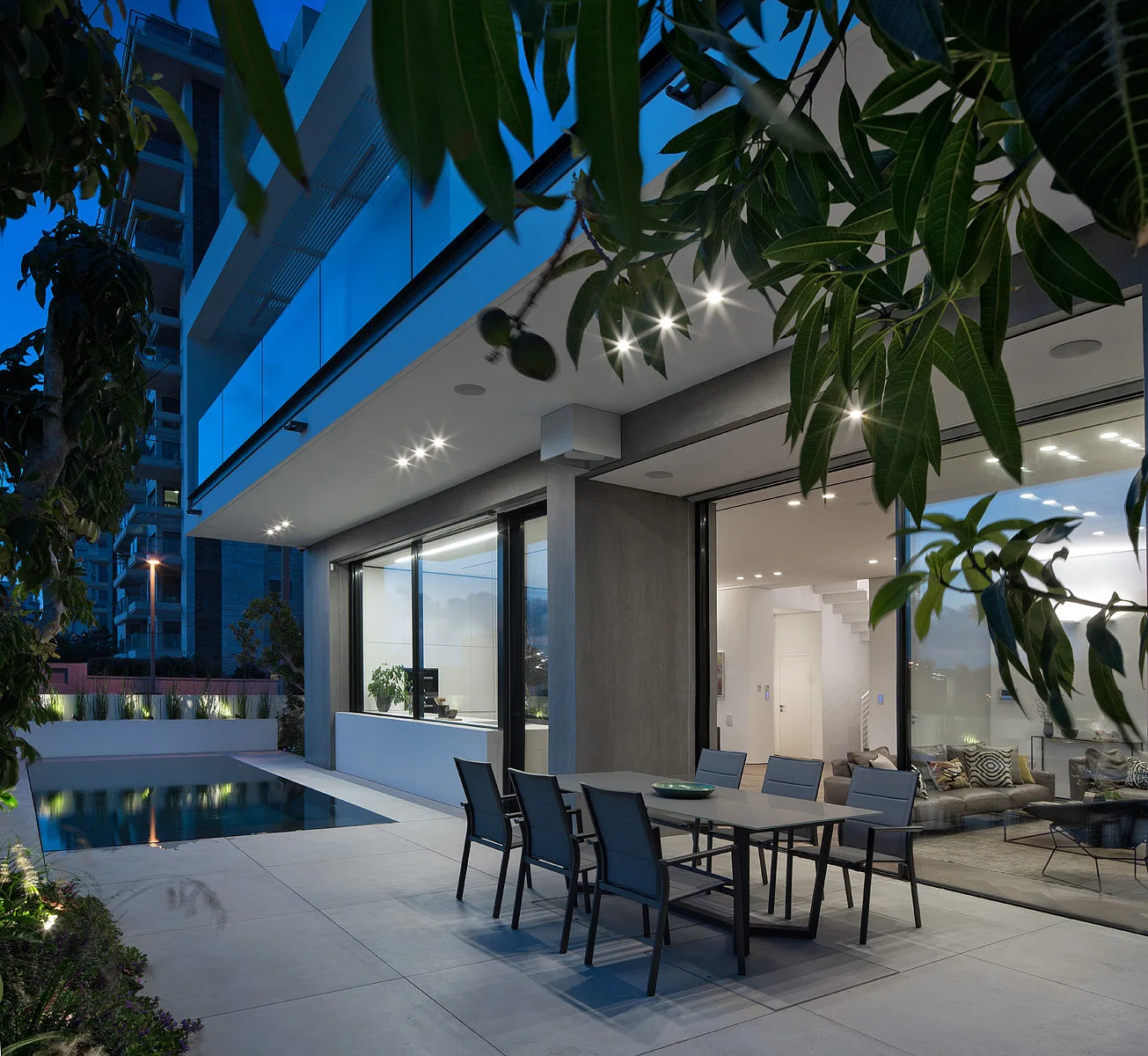 A modern grey patio features a metal dining set next to a rectangular swimming pool under recessed ceiling lights at dusk.