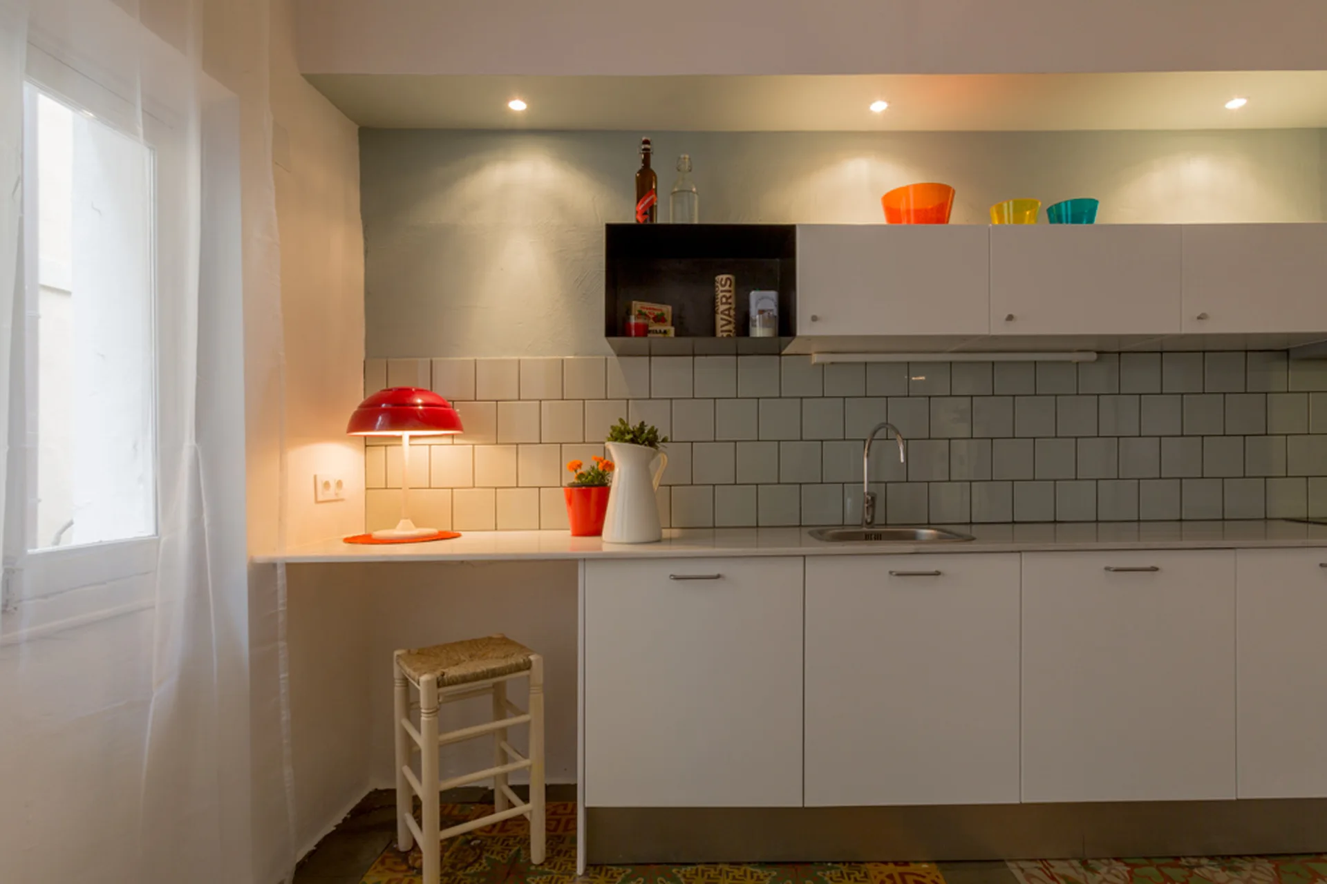 A minimalist kitchen features a white countertop with a red desk lamp, a light grey tile backsplash, white cabinets, and a woven wooden stool.