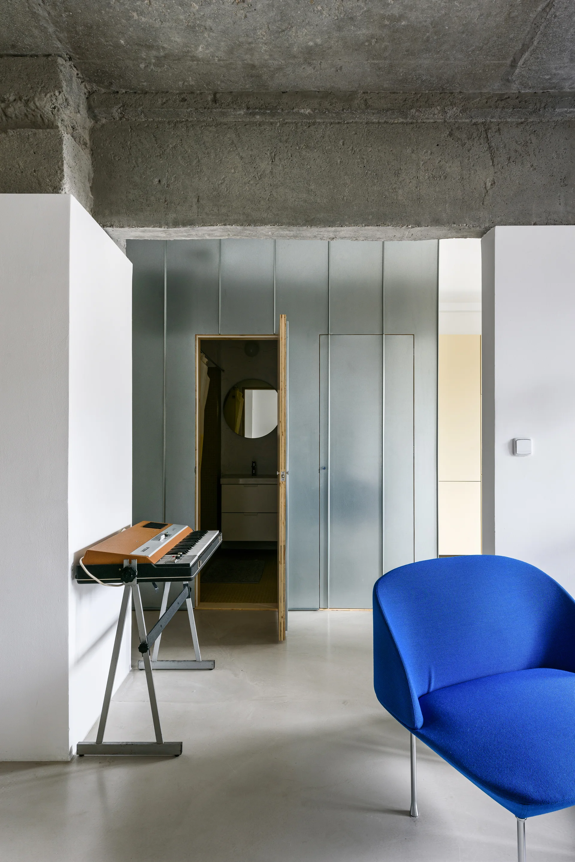 A modern room with a raw concrete ceiling, a galvanized metal paneled partition wall, a bright blue armchair, and a keyboard on a stand.