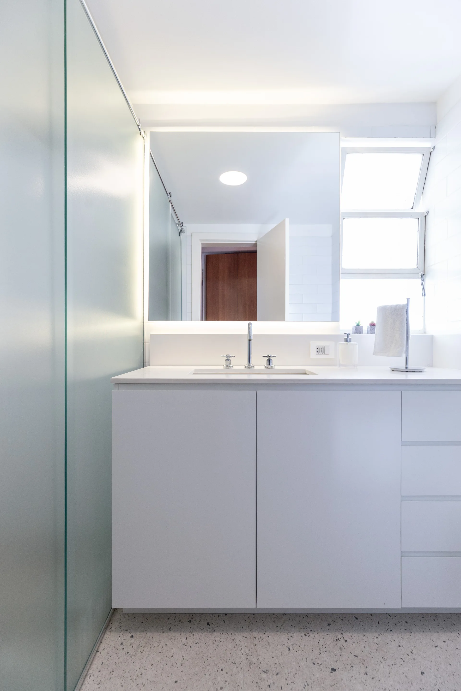 A modern white bathroom vanity with a chrome faucet, a large backlit mirror, and grey terrazzo flooring.