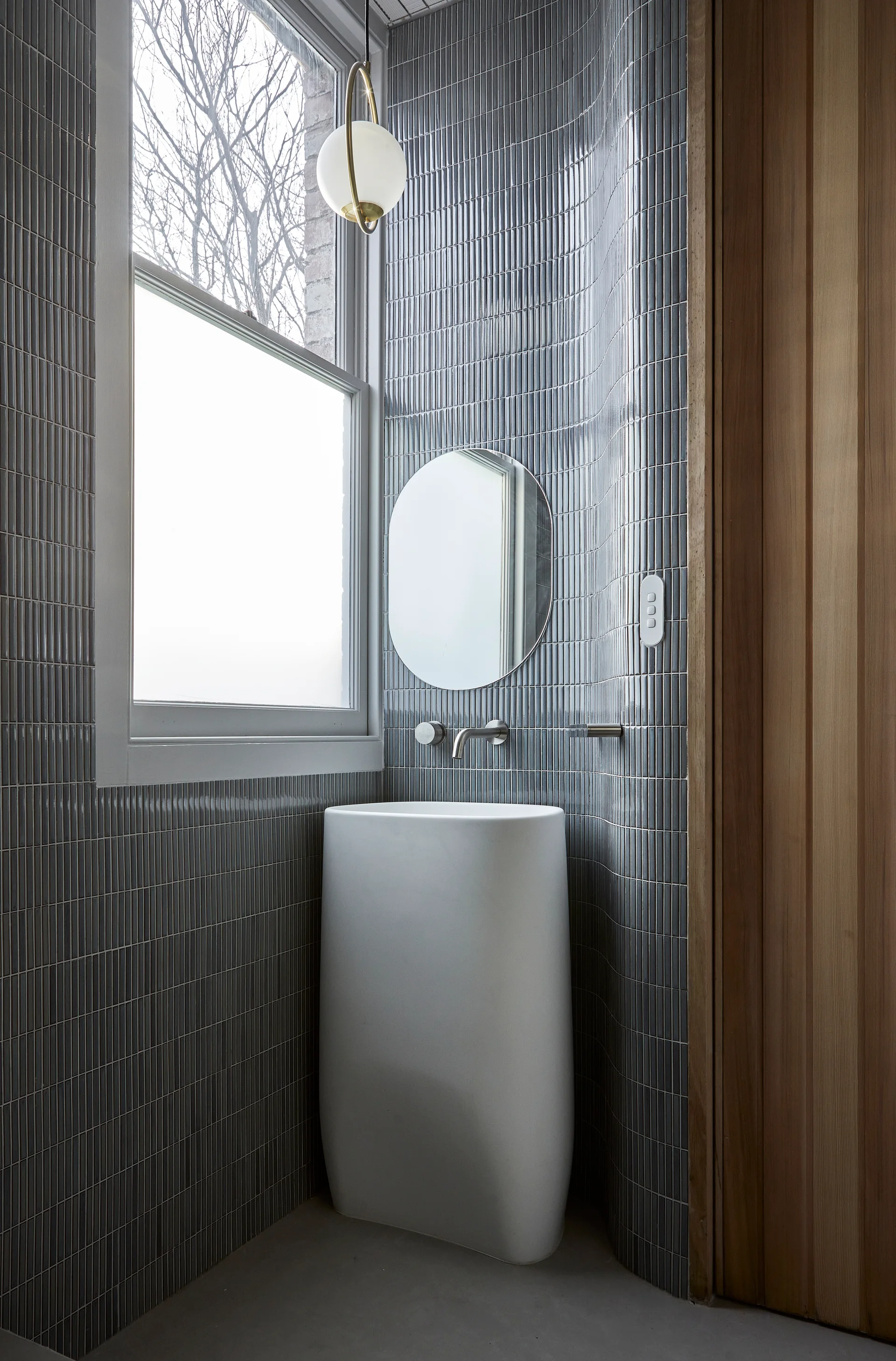 A minimalist powder room features a white freestanding pedestal sink beneath a round mirror on a curved wall with grey vertical tiles.