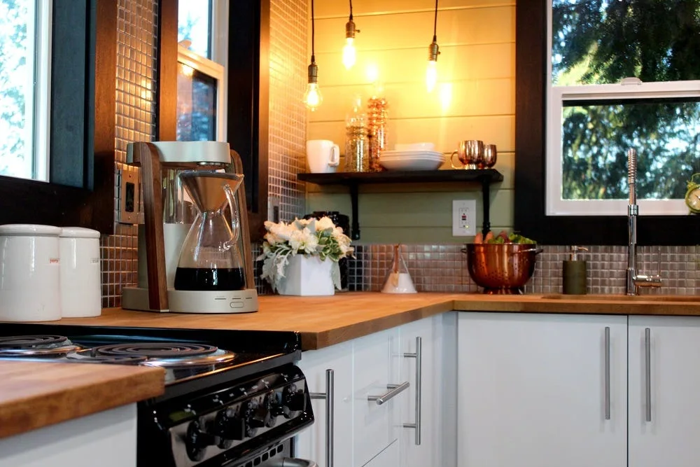 A kitchen counter with a butcher block top features white cabinets, a small glass mosaic tile backsplash, and hanging exposed bulb pendant lights.
