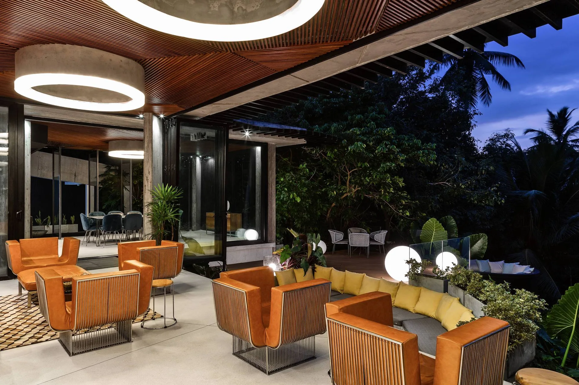 An outdoor patio features orange armchairs with metal wire bases, a built-in sofa with yellow cushions, and glowing sphere floor lamps at twilight.