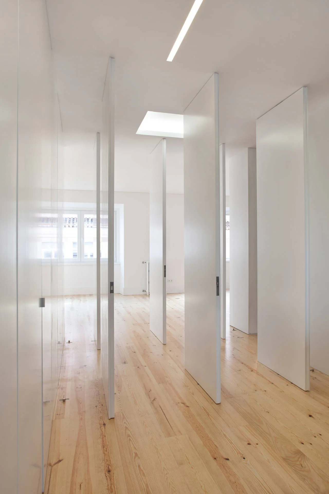 A long minimalist hallway features several tall white doors opening along a light wood plank floor beneath a skylight.