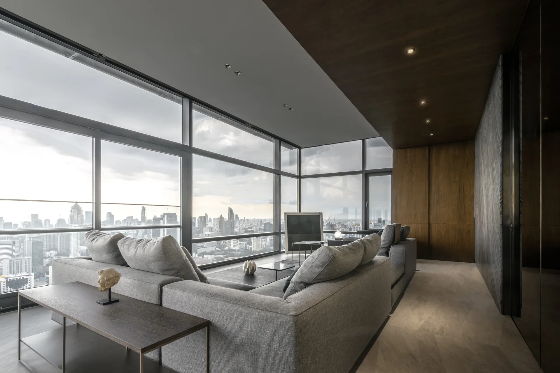 A modern high-rise living room features a large grey sectional sofa, floor-to-ceiling windows with city skyline views, and dark wood wall paneling.