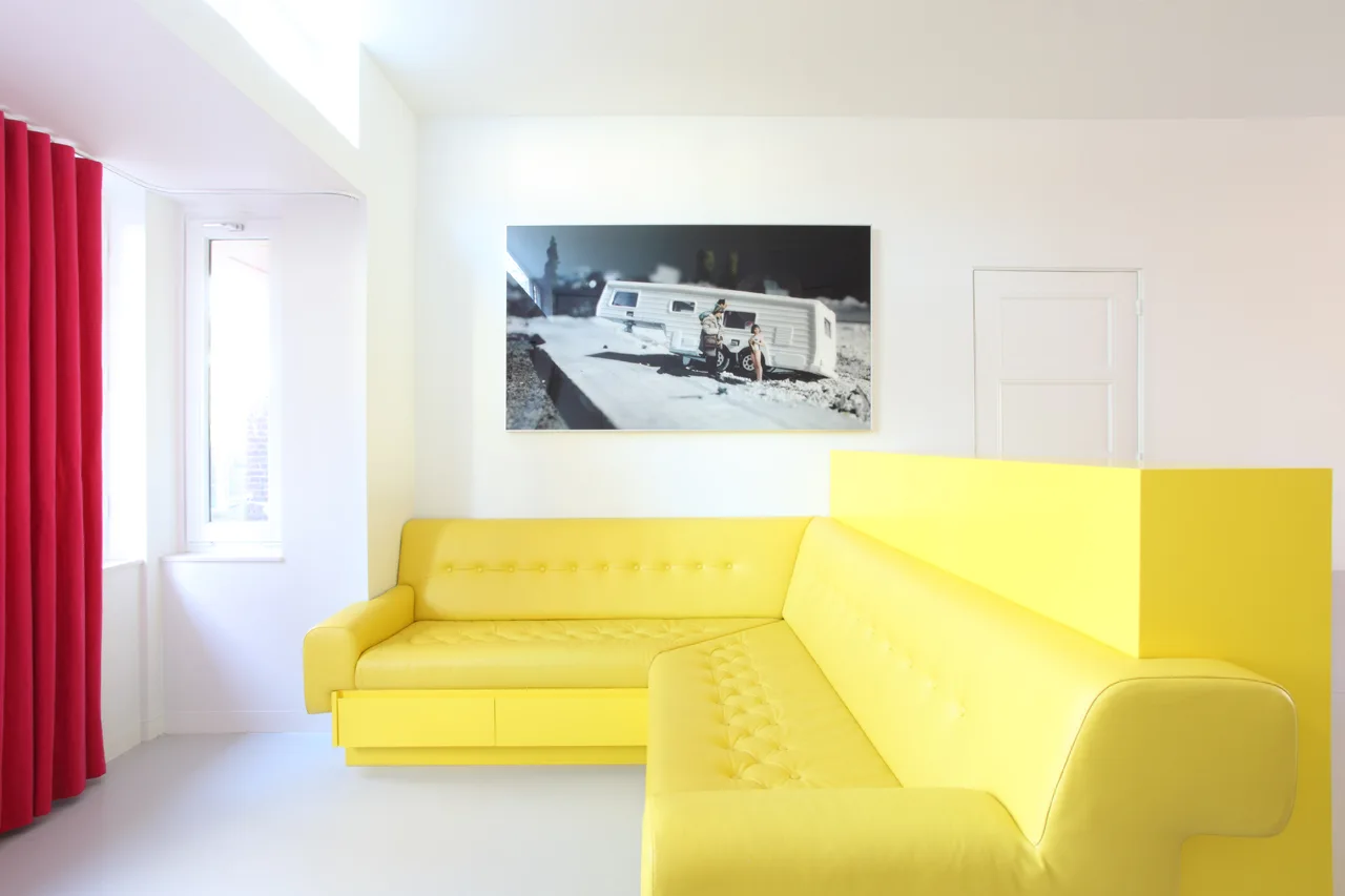 A bright yellow tufted corner sofa sits against stark white walls next to a long red curtain and a black and white framed wall art piece.