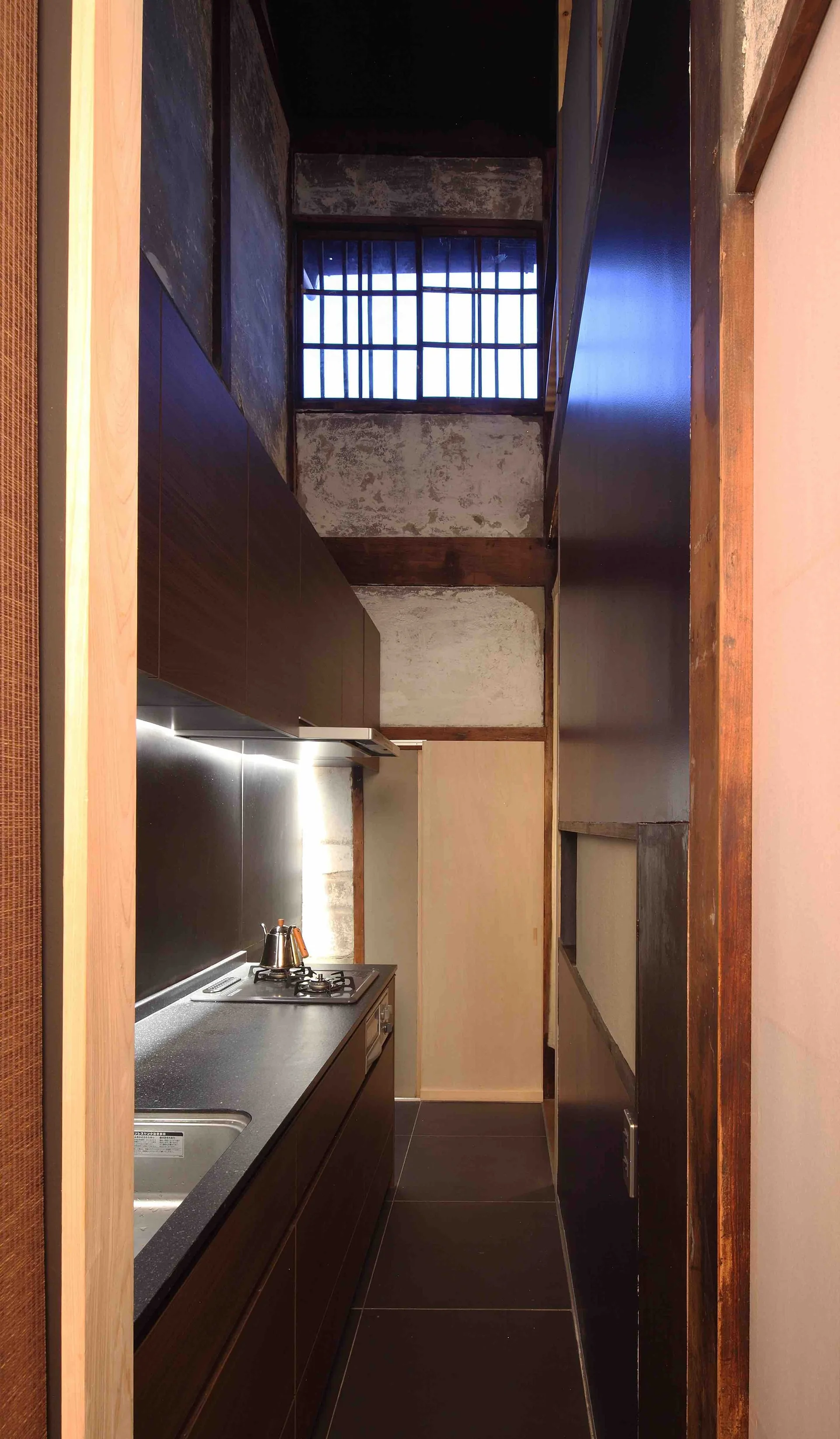 A narrow galley kitchen features dark wood cabinets, a black countertop with under-cabinet lighting, and weathered plaster walls with exposed timber beams.