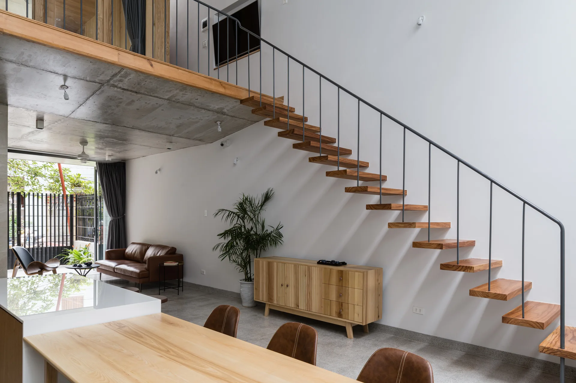 A modern open-plan living area features a floating wooden staircase, an exposed concrete ceiling, a slatted wood sideboard, and brown leather furniture.
