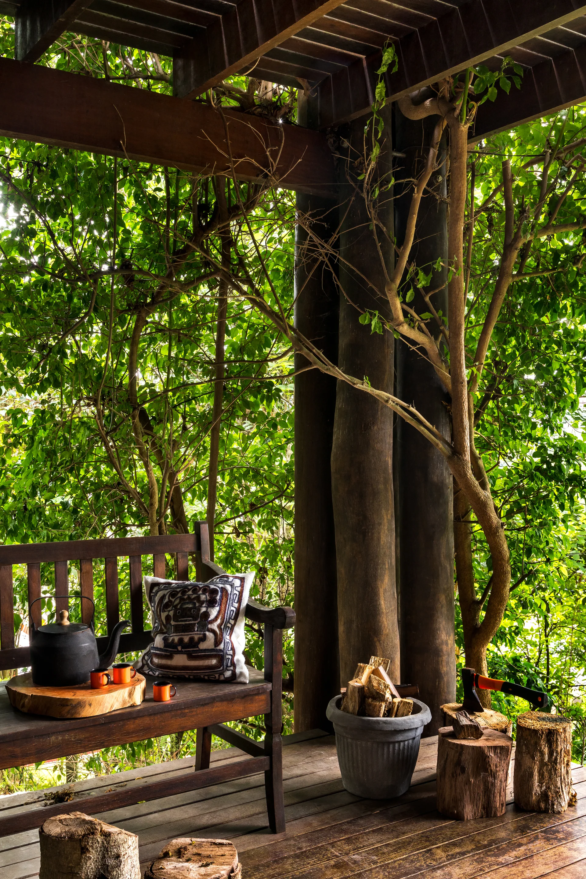 A rustic wooden bench with a patterned pillow sits on a deck under a slatted pergola, surrounded by dense green trees.