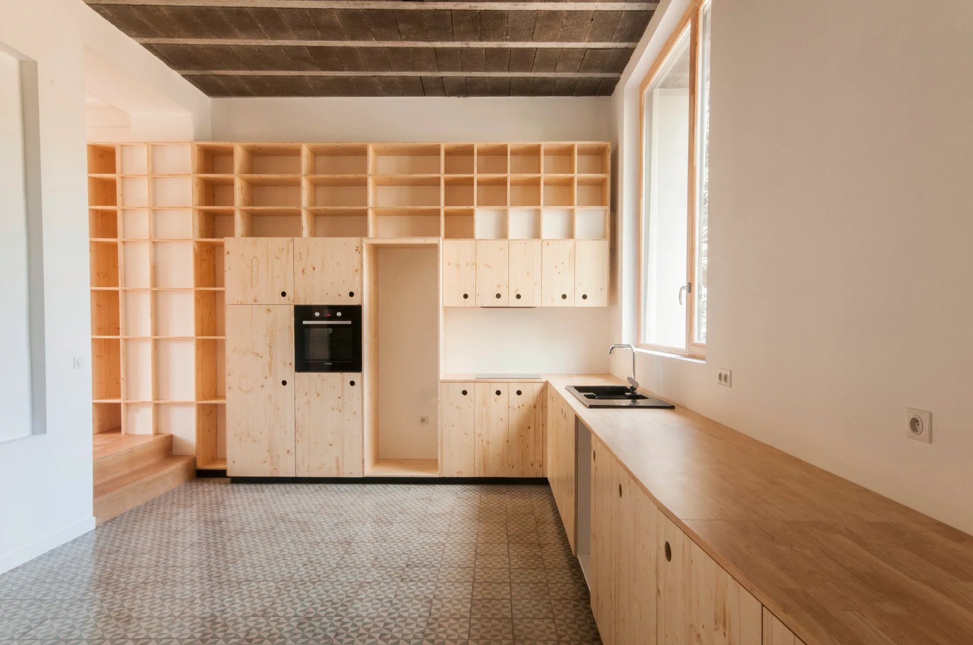 A minimalist kitchen featuring light wood cabinets, built-in grid shelving, a black sink, and geometric-patterned floor tiles under a concrete ceiling.