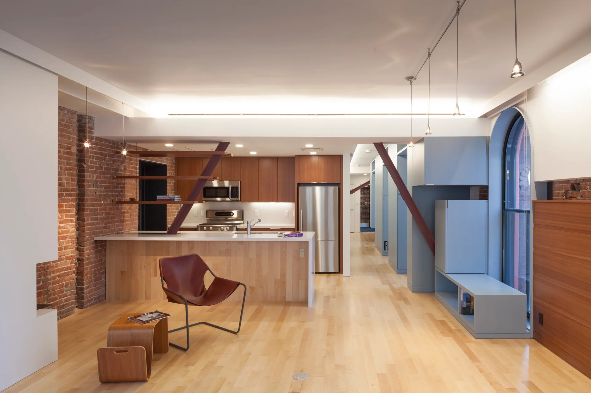 An open loft kitchen and living area with blonde wood floors, exposed brick walls, a wooden island, and custom blue cabinetry near an arched window.