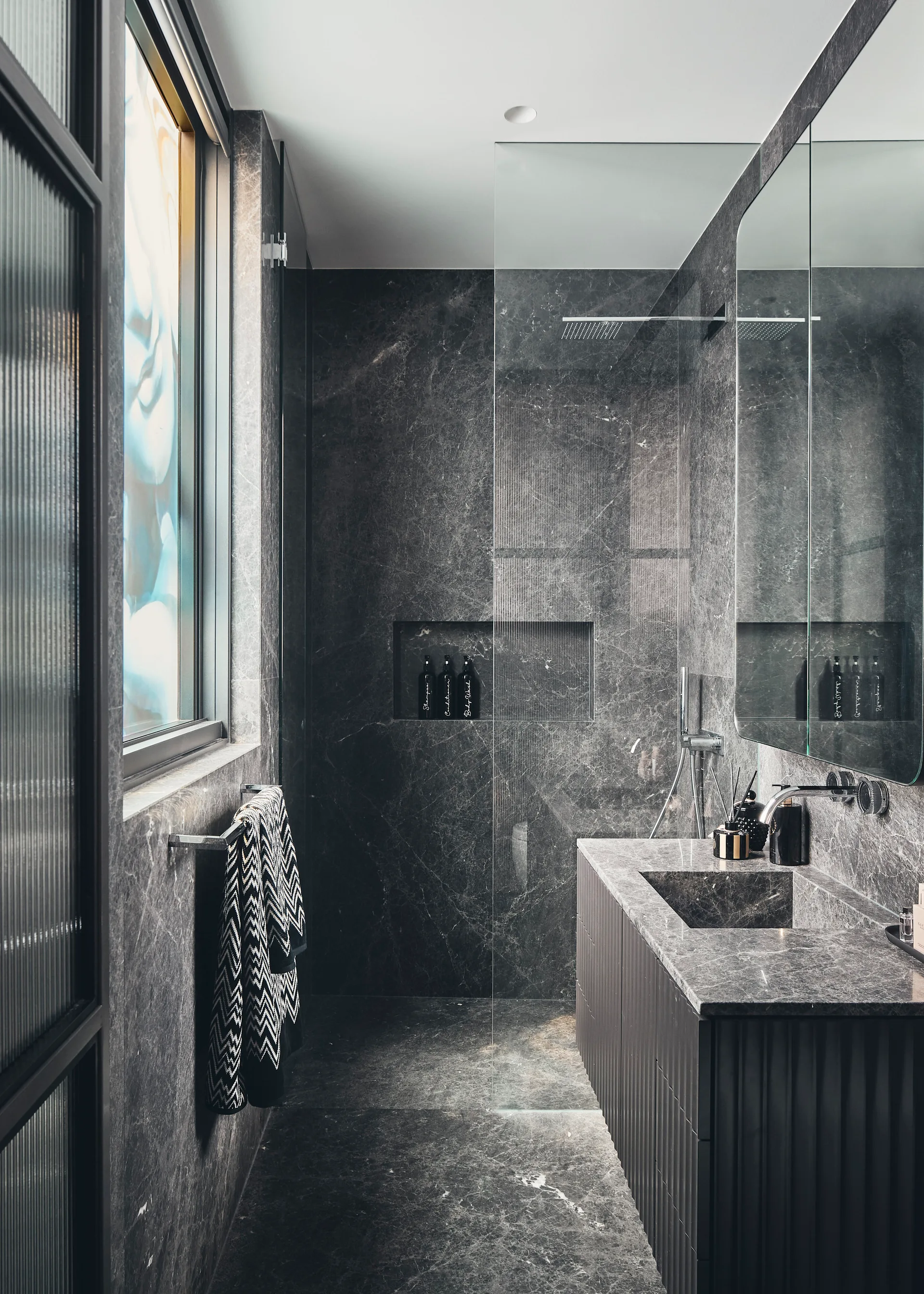 A contemporary bathroom lined with dark gray veined marble features a glass shower panel and a fluted vanity with an integrated stone sink.