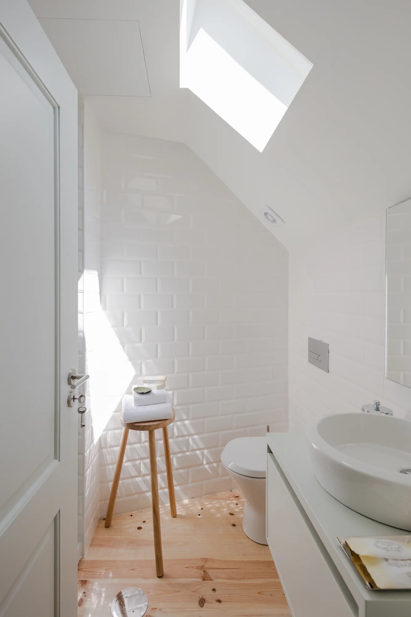 A small attic bathroom features white subway tiled walls, a wooden tripod stool, and a skylight casting natural light onto the light wood floor.