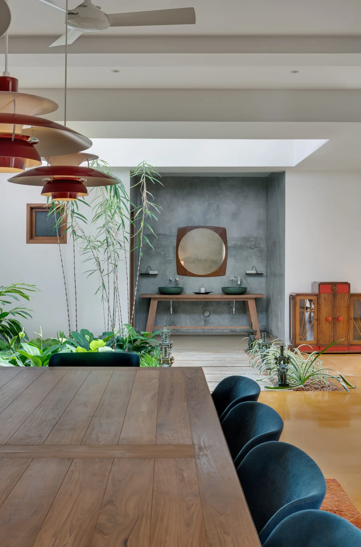 An open dining area with a wooden table and blue chairs overlooks an indoor garden path leading to a concrete-finished vanity alcove.