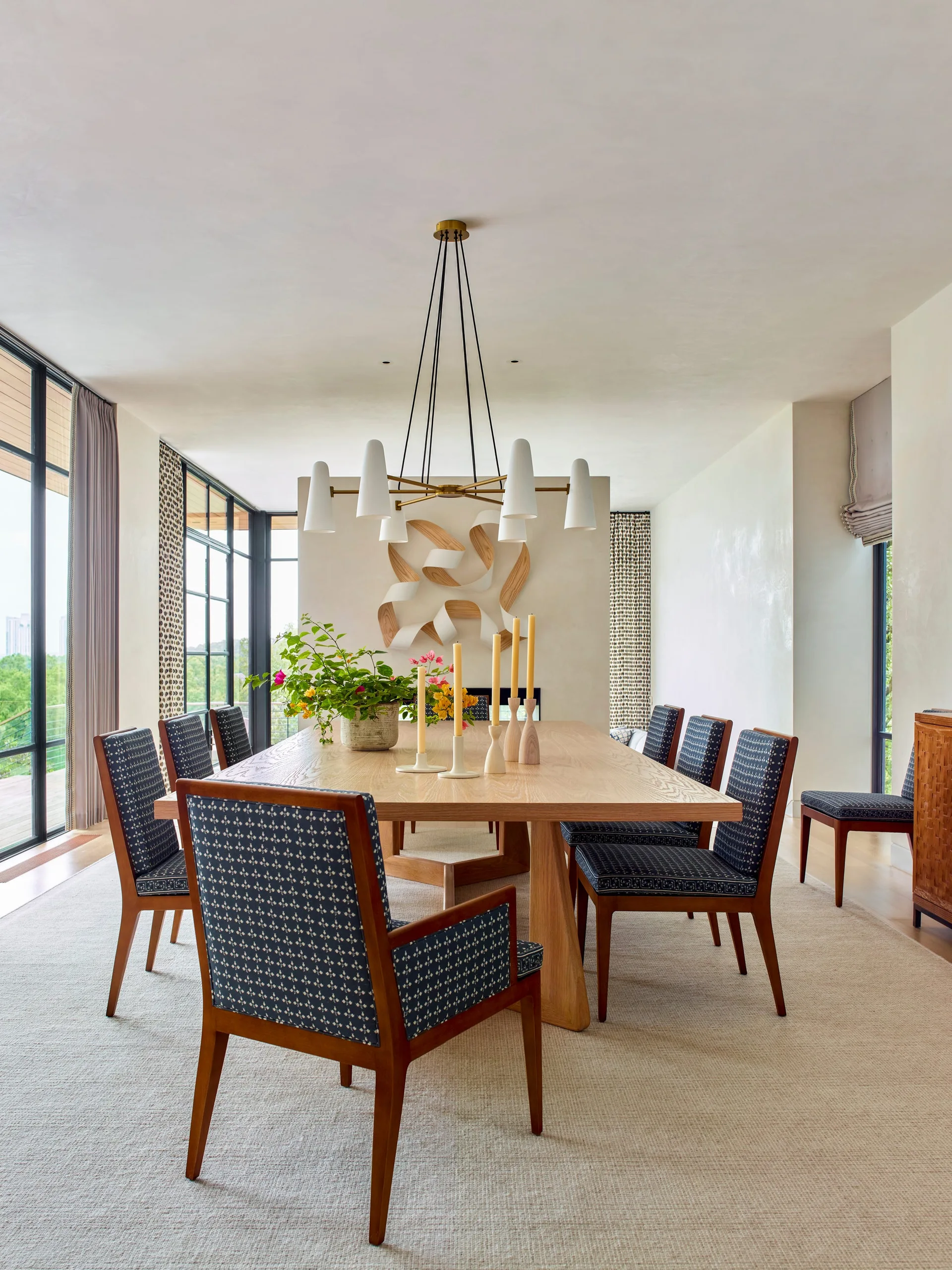 A contemporary dining room features a light oak table surrounded by navy patterned chairs under a white multi-arm chandelier.