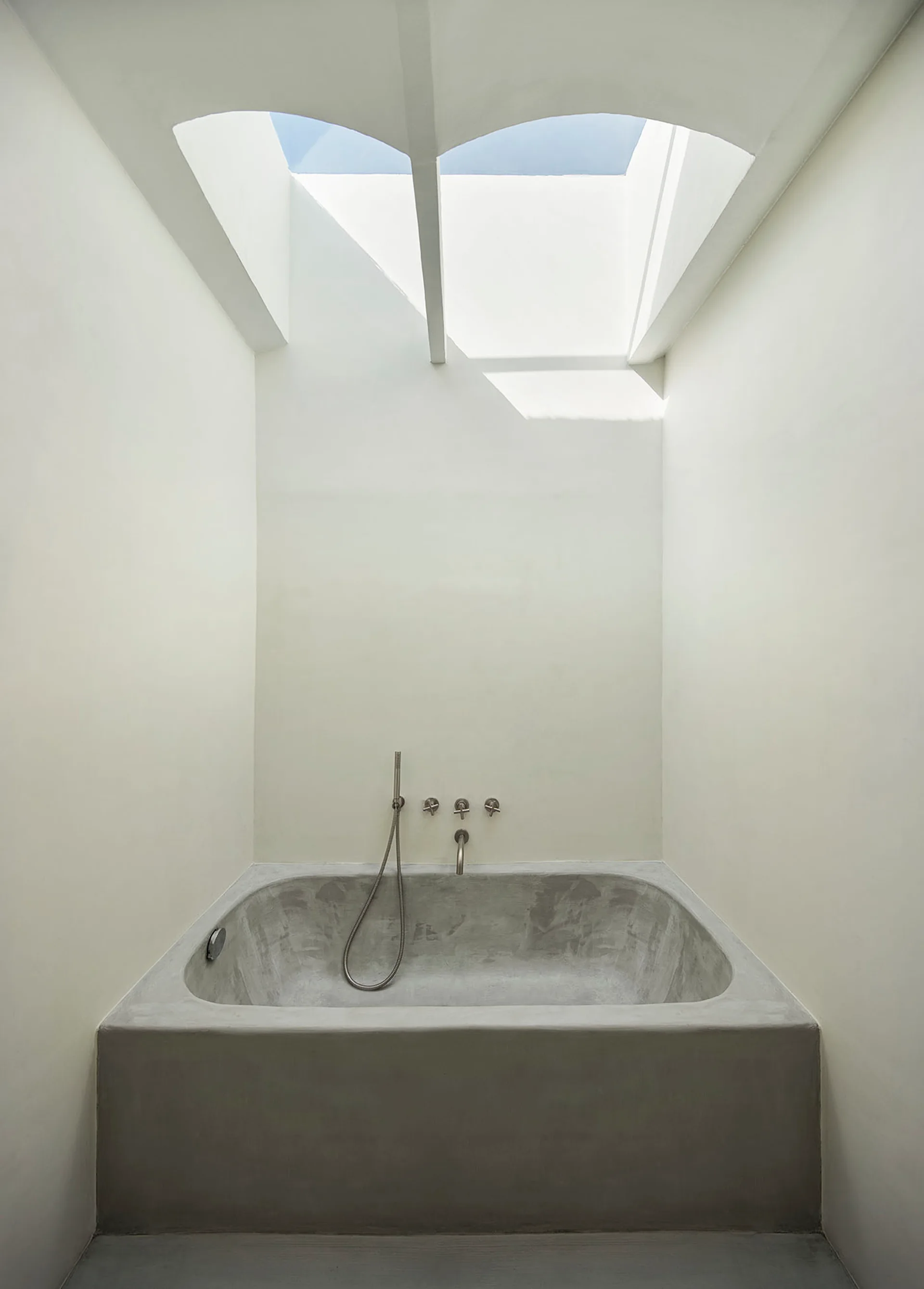 A minimalist grey concrete bathtub is built into off-white walls beneath an arched ceiling skylight that lets in natural light.