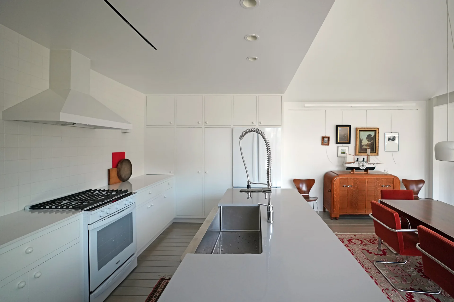 A minimalist white kitchen features a grey island with a chrome faucet, white cabinetry, and an adjacent dining area with red chairs.